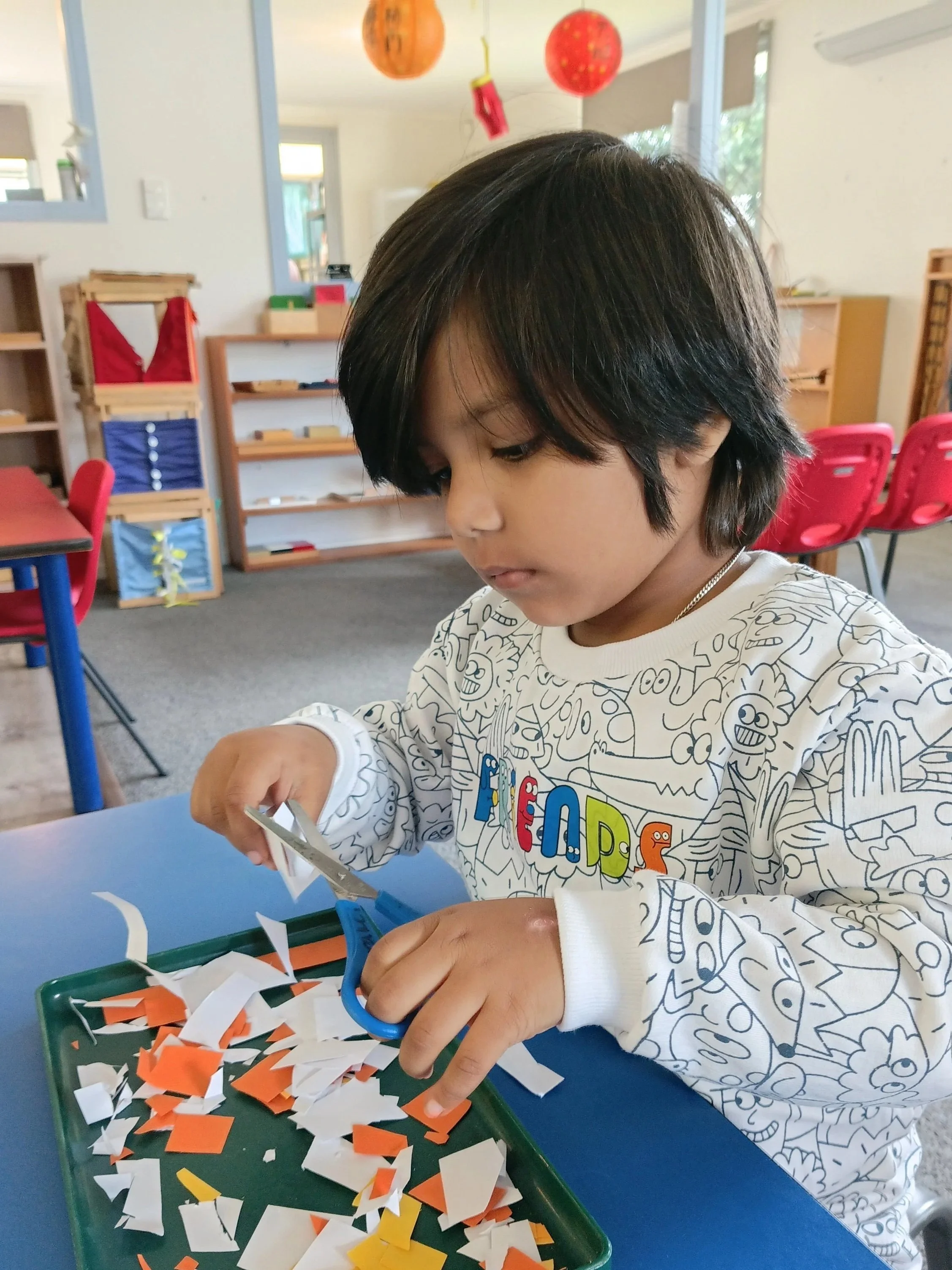 A young boy with black hair wearing a white sweater with colorful lettering, sitting at a table, cutting small pieces of paper with scissors in a classroom setting.