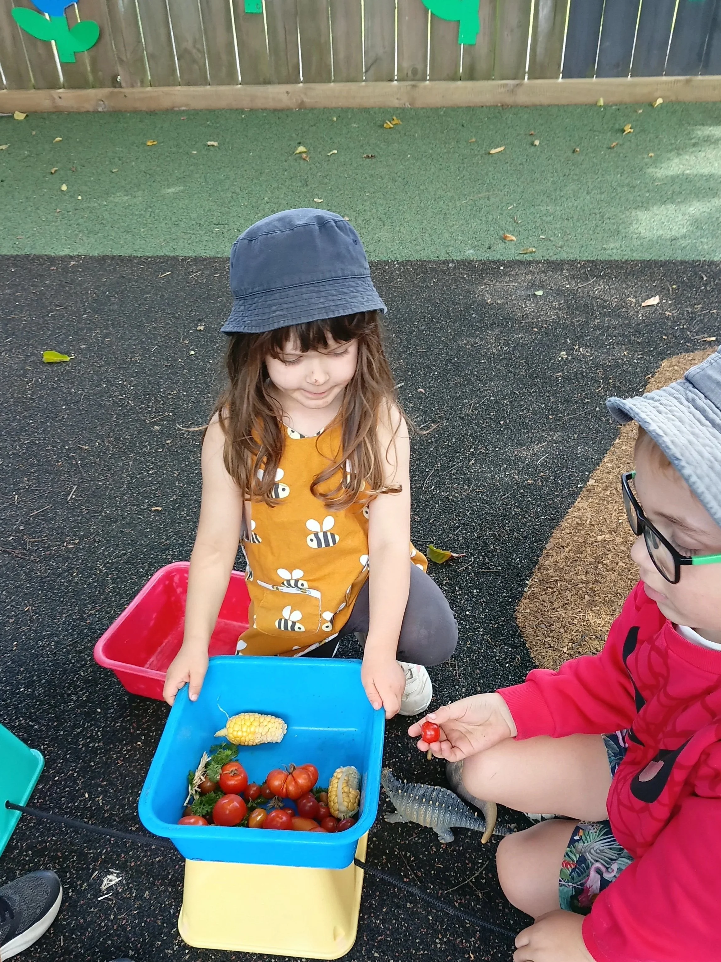 Two children, a girl and a boy, outdoors with containers of toy vegetables. The girl holds a toy tomato, and the boy holds a small red object, possibly a toy vegetable, as they play pretend with toy garden produce.