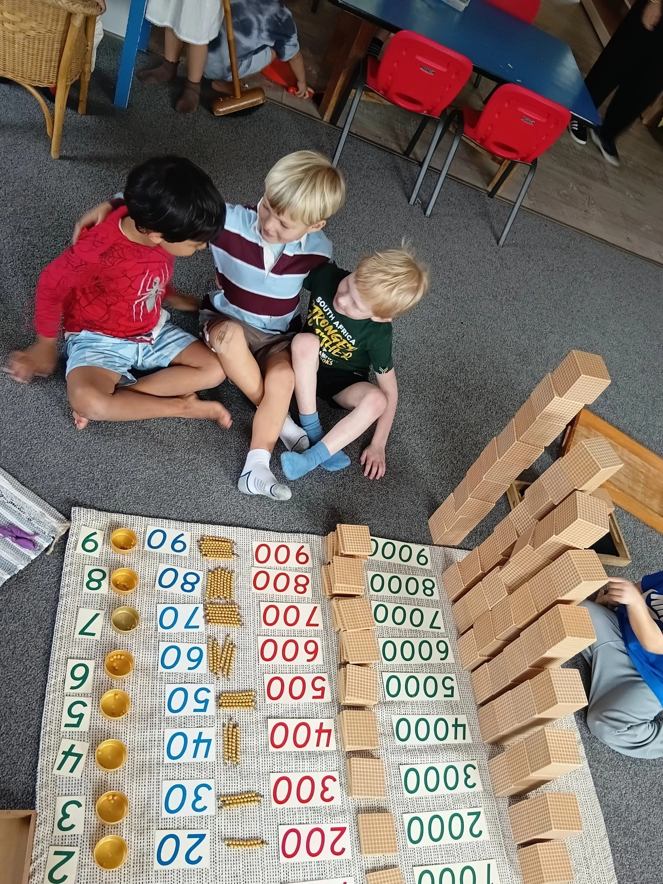 Three children sitting on the carpet in a classroom, engaging in a math activity with number tiles, small bowls, and building blocks arranged on a board.