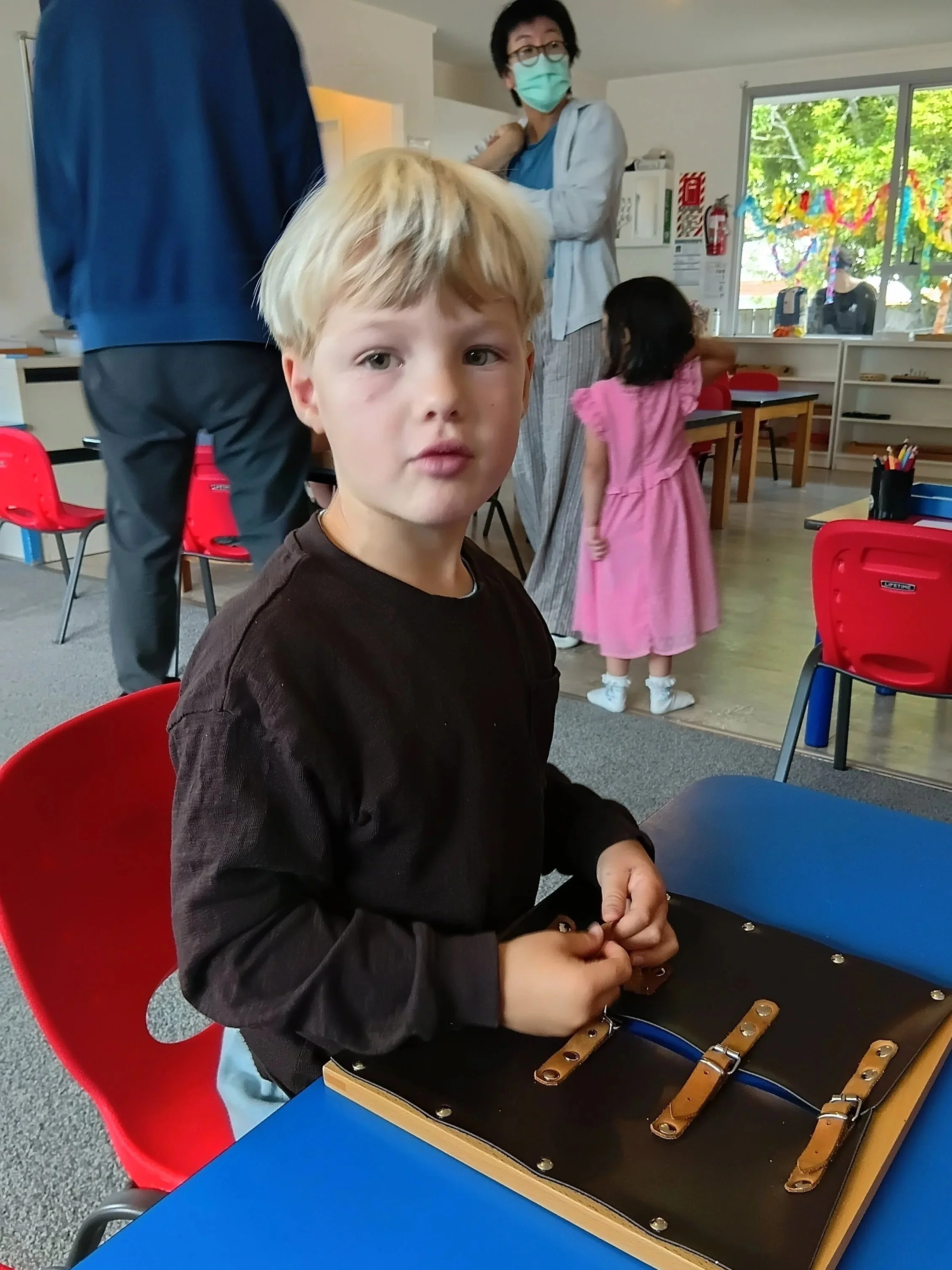 A young boy with blonde hair sits at a blue table in a classroom, looking directly at the camera. He appears to be working on a leather crafting project with leather straps and rings. In the background, a woman wearing a mask and glasses, a blue shir