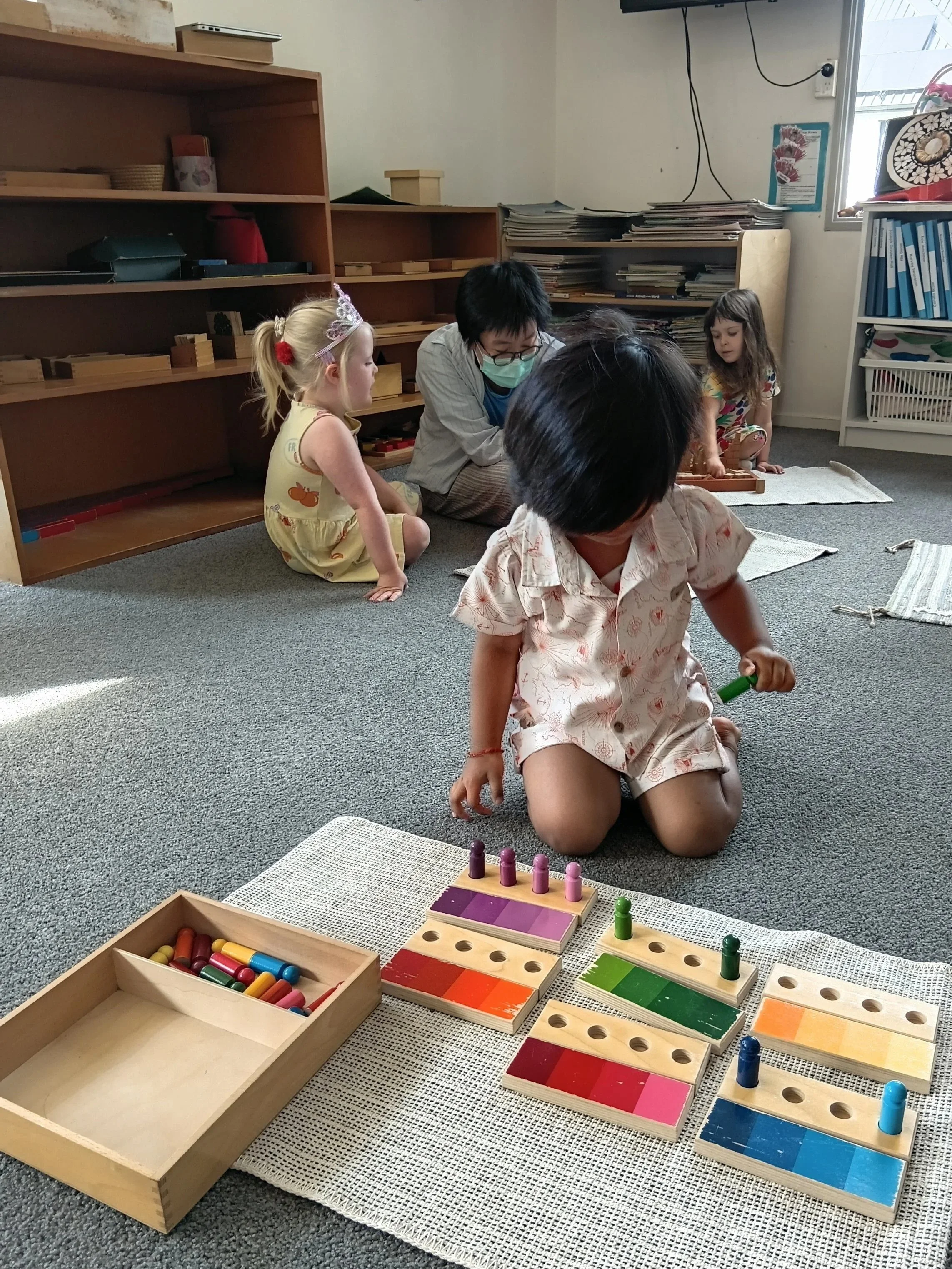 Children playing with colorful wooden stacking and sorting toys on a white rug in a classroom setting, with an adult supervising in the background.