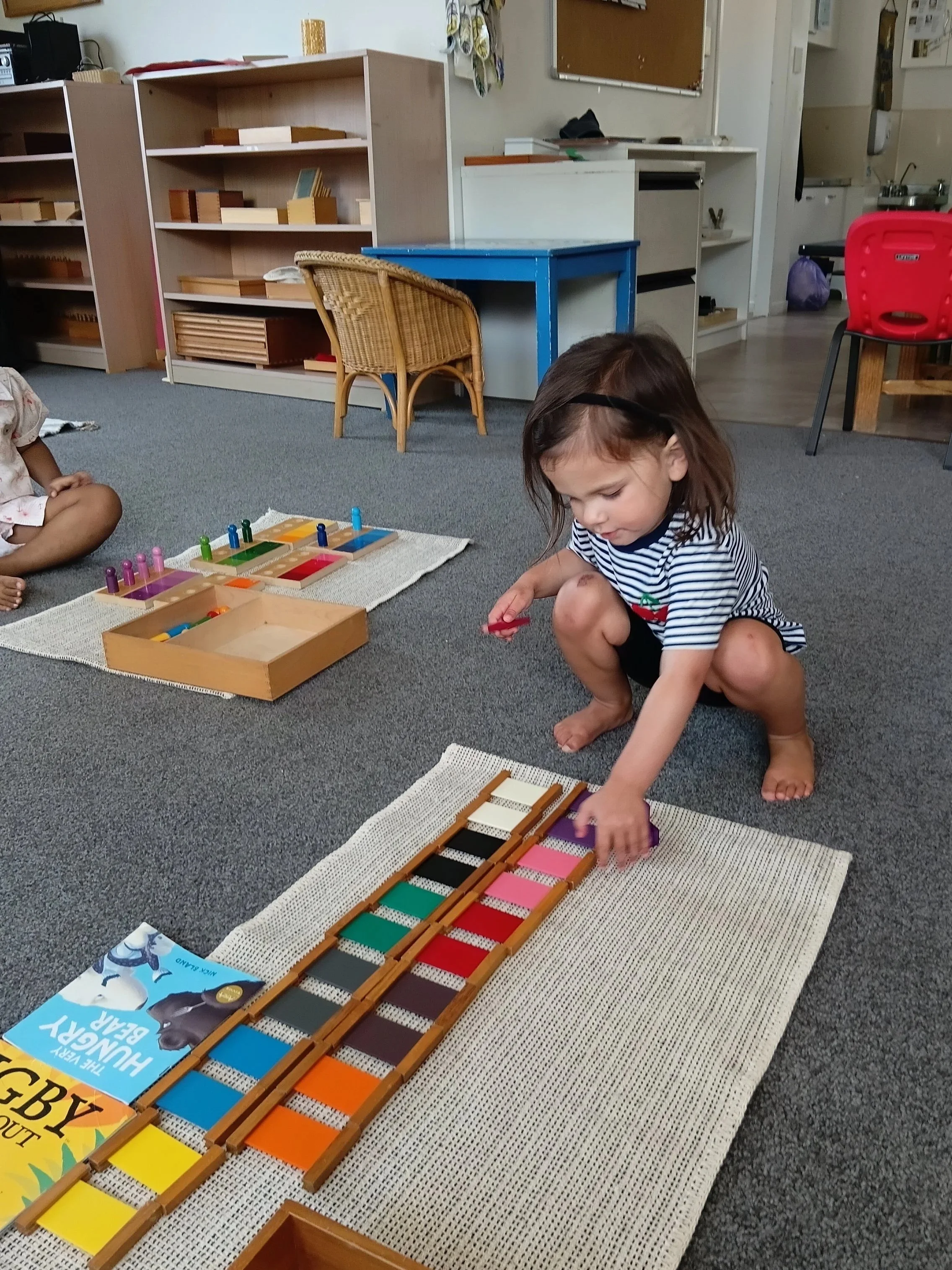 Child crouching on carpeted floor playing with large colorful xylophone or musical instrument in classroom.