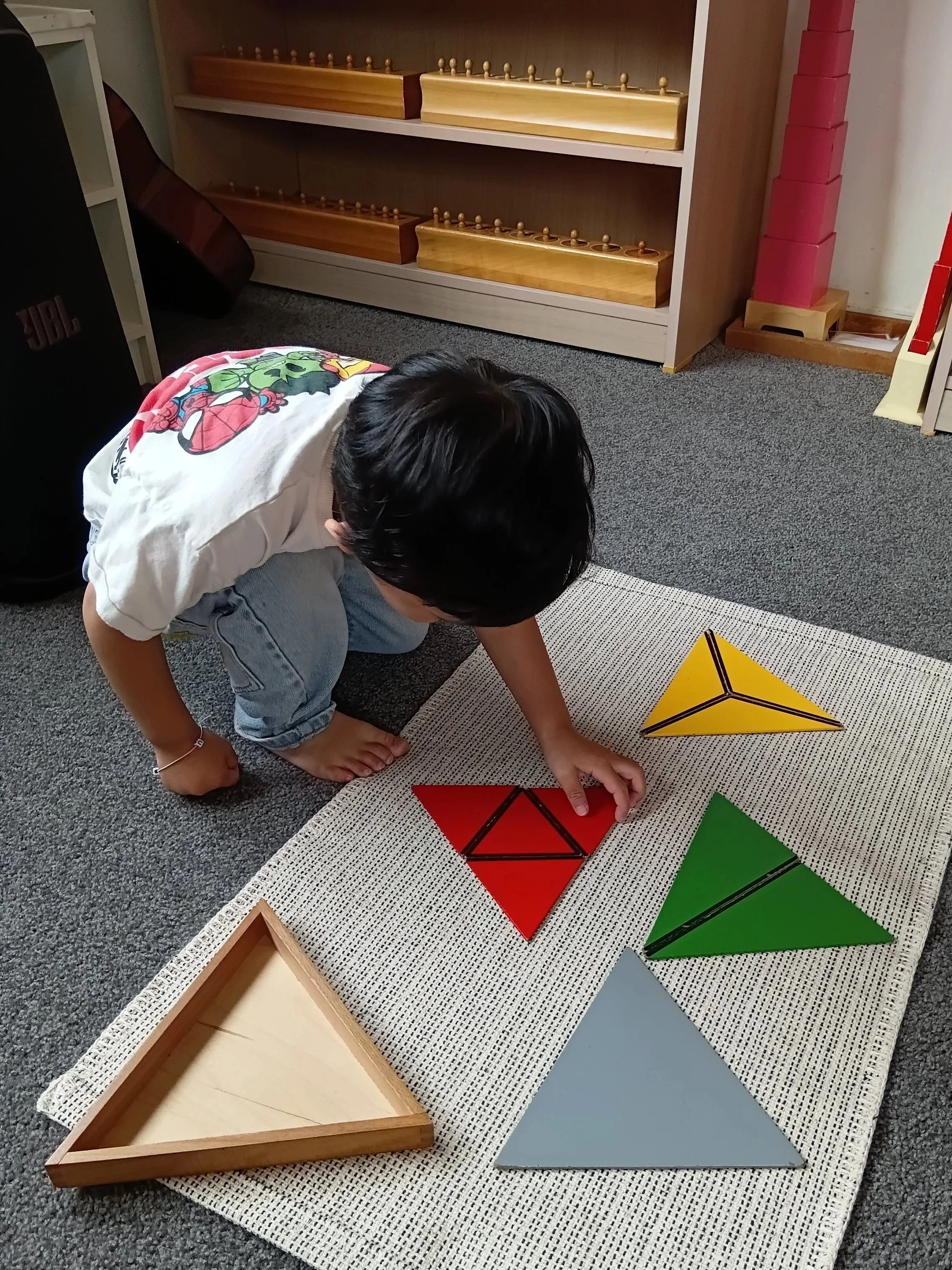 A young child is crouched on a carpeted floor, playing with colorful geometric puzzles that are in the shapes of triangles and a square, on a white textured mat.