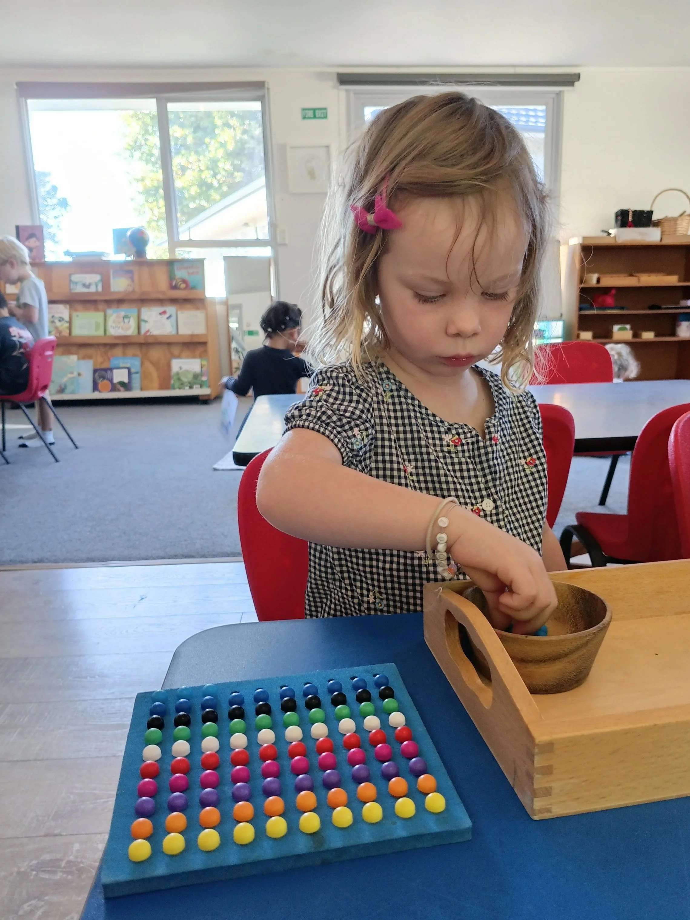 Young girl with a pink bow in her hair, playing with an abacus and a wooden bowl at a table in a classroom.