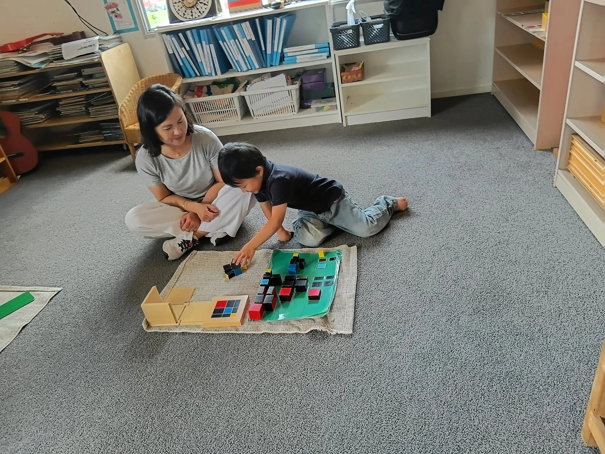 A woman and a young boy playing with colorful blocks on a small rug in a classroom setting.