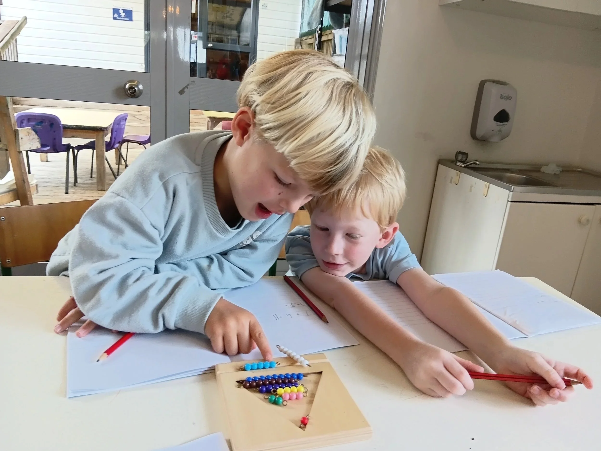 Two young boys sitting at a table working on a school activity with a multiplication setup and colored beads. The boy on the left points at the beads while the boy on the right holds a red pencil and looks on.