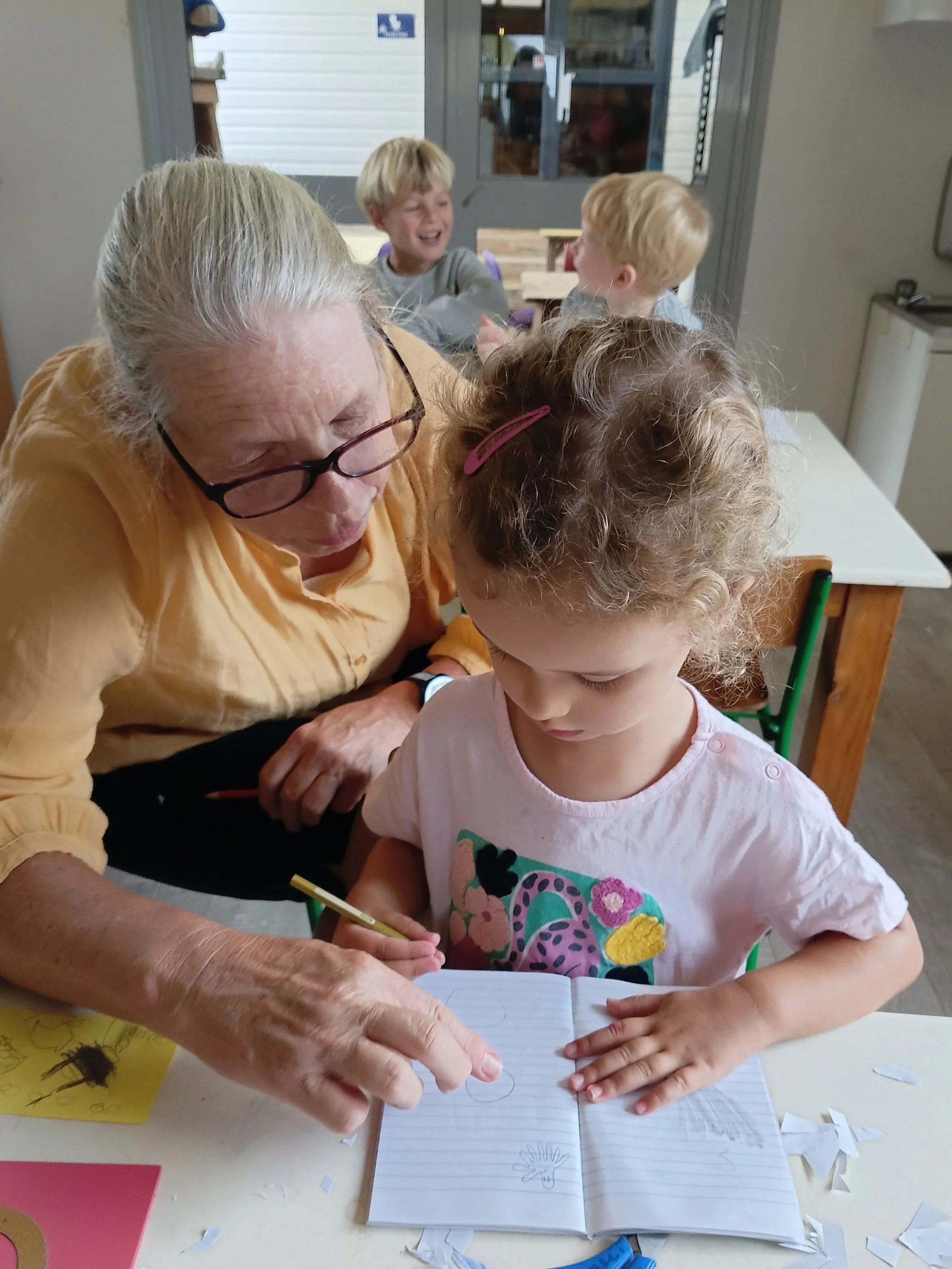 An elderly woman helping a young girl draw in a notebook at a classroom table, with three children in the background laughing.