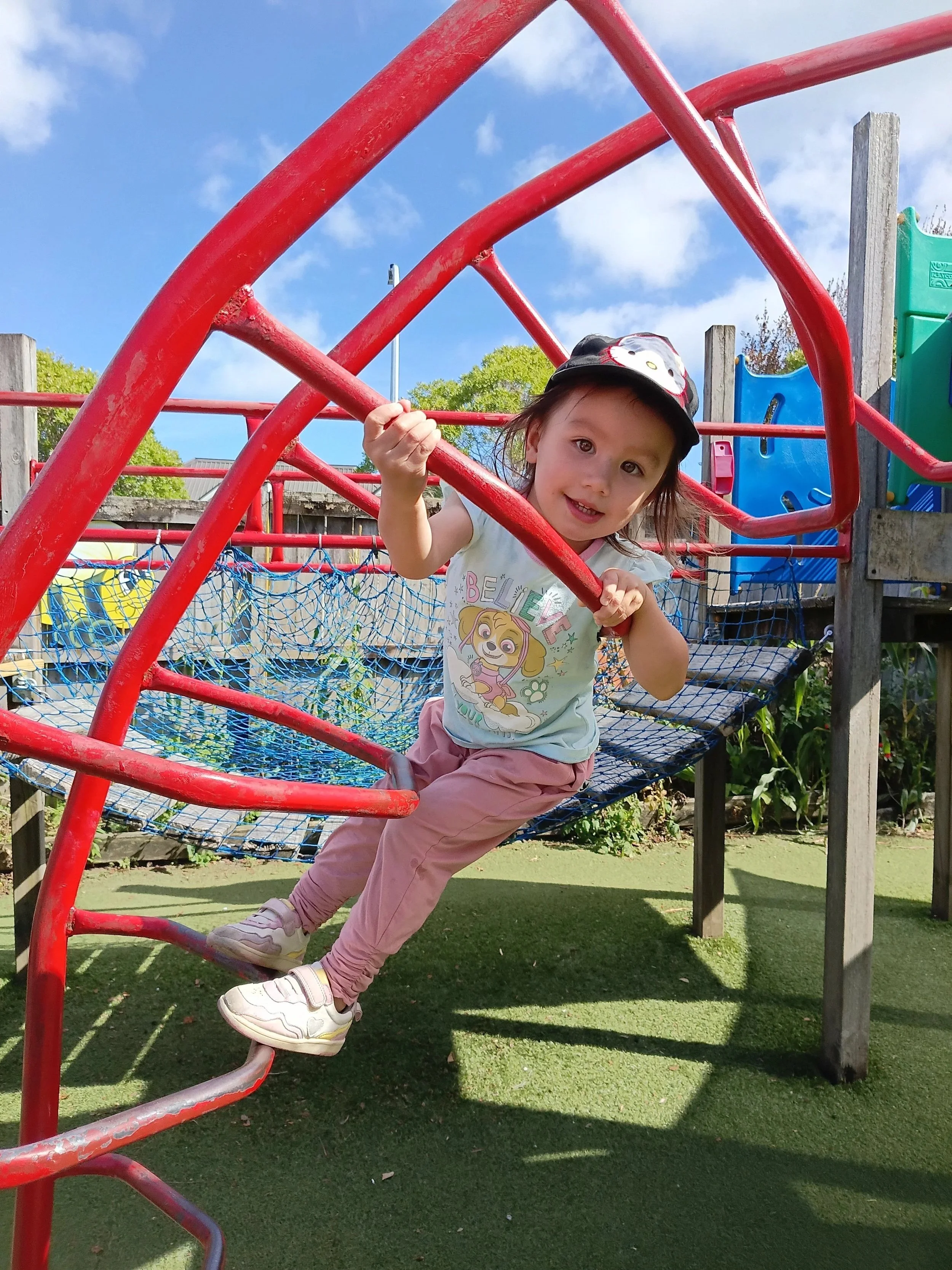 Young girl wearing a cartoon character t-shirt, pink pants, and white sneakers playing on a red jungle gym at a playground, under a clear blue sky.
