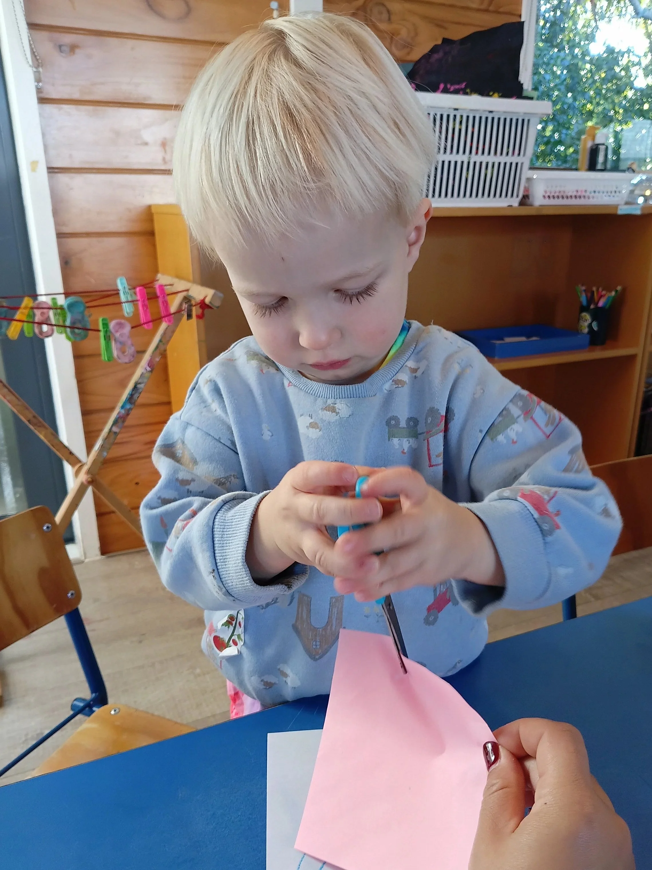 A young boy with blonde hair and a light blue shirt featuring a train pattern is sitting at a table, focused on a craft activity involving pink paper and a sewing needle held by an adult hand. The setting appears to be a classroom or playroom, with w