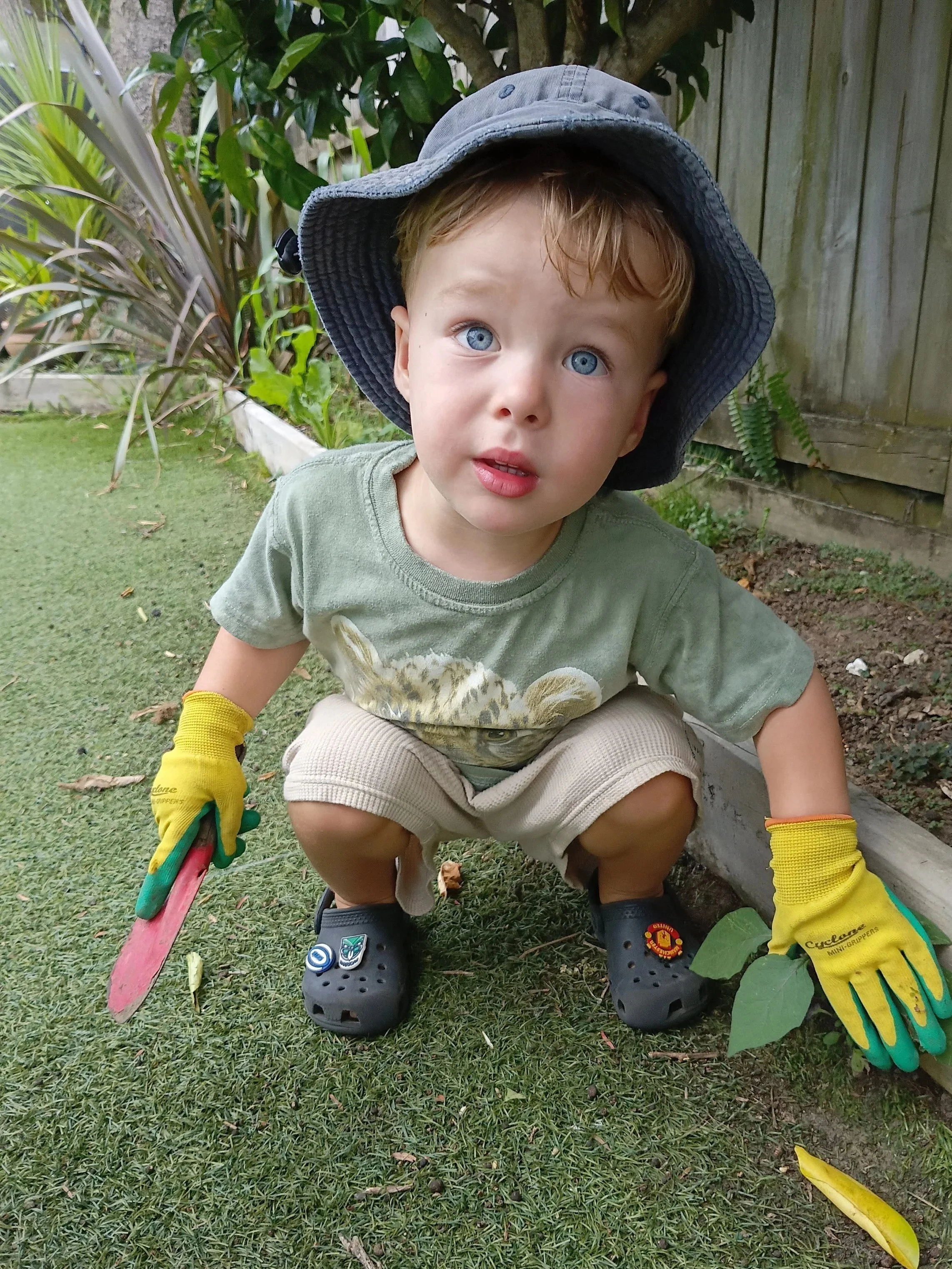 Young boy with blonde hair and blue eyes is squatting outdoors near a garden bed, wearing a wide-brimmed gray hat, yellow gardening gloves, and Crocs. He holds a red gardening tool in his right hand and is looking up at the camera.