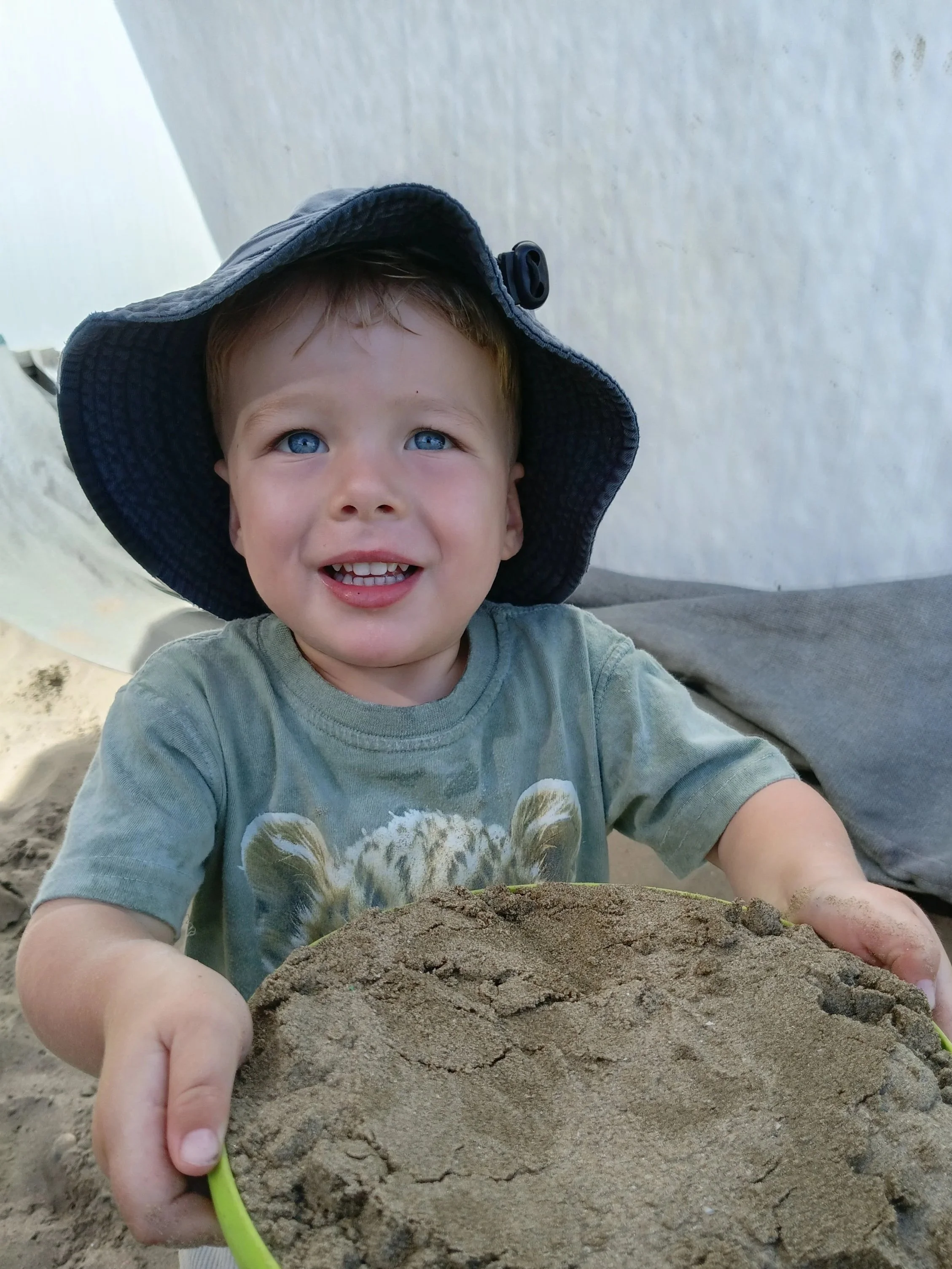 A young boy wearing a blue sun hat and a green t-shirt with a tiger print, smiling and holding a green bowl filled with sand at the beach.