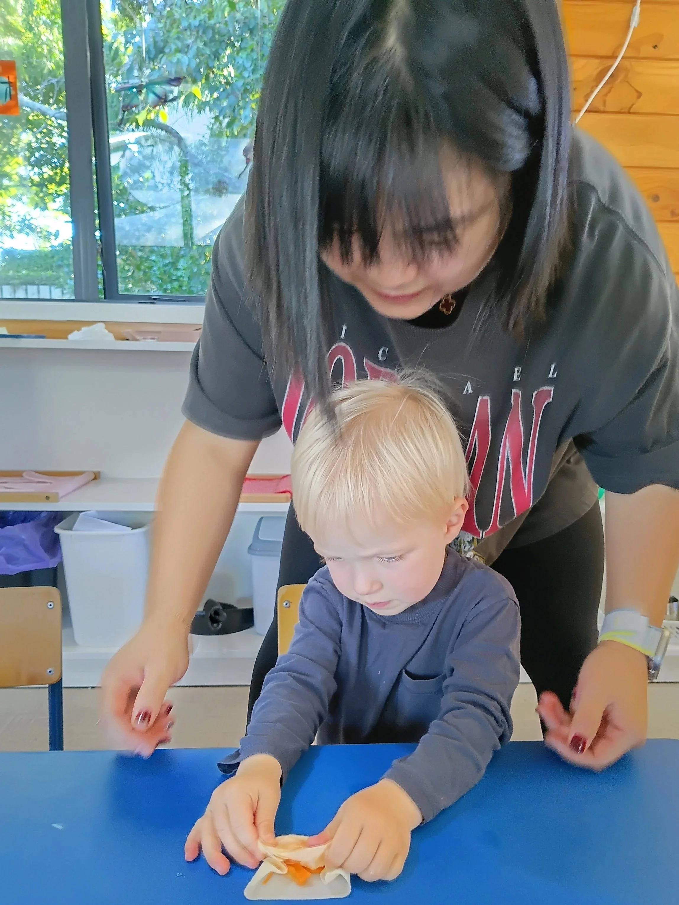 A woman with black hair and glasses assisting a young blonde boy with a craft project at a blue table in a classroom.