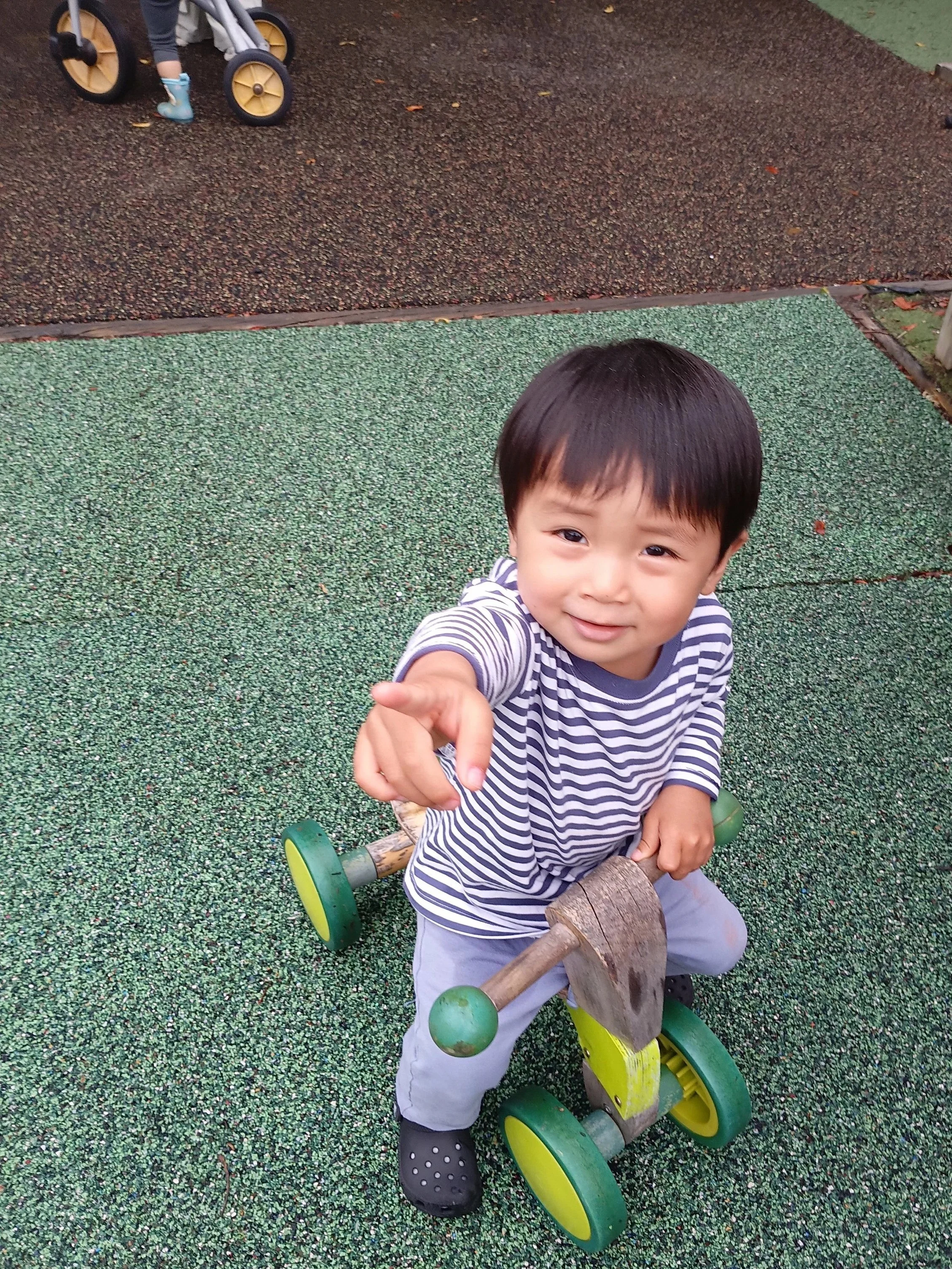A young boy sitting on a wooden and green tricycle outdoors on a rubberized play surface, pointing at the camera, wearing a striped shirt and gray pants.