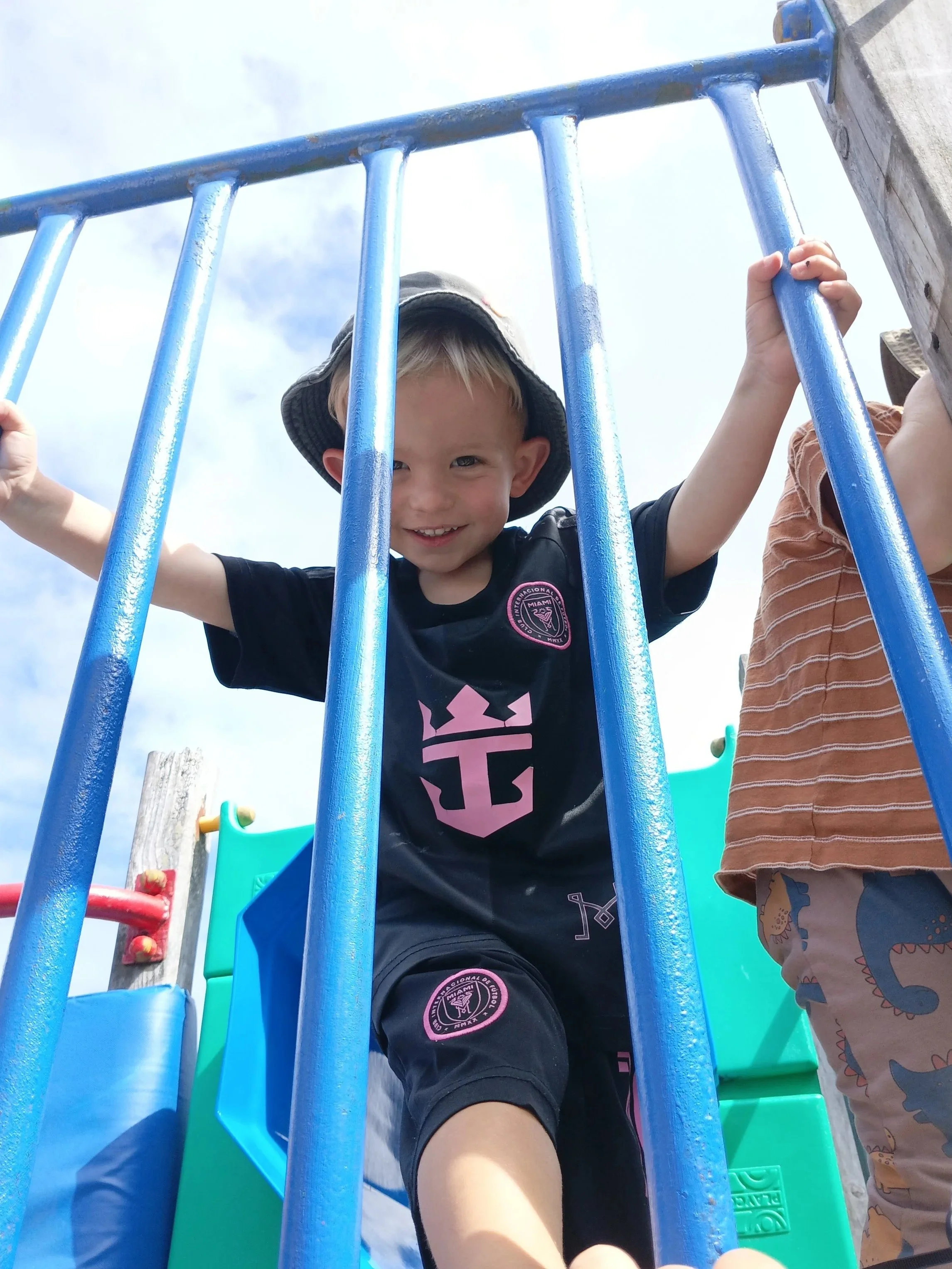 A young boy with a bucket hat smiling while climbing a colorful playground structure, holding onto blue bars, with another child's arm visible nearby, under a partly cloudy sky.