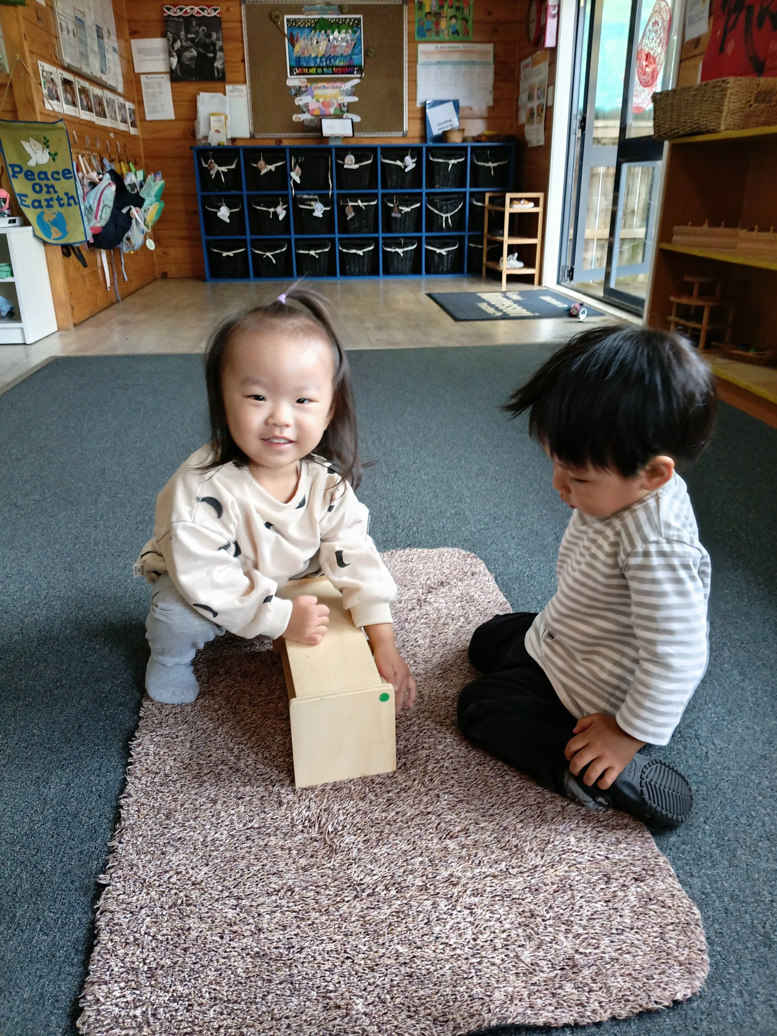 Two preschool children, a girl and a boy, sitting on a small rug in a classroom. The girl is smiling and holding a small wooden box, while the boy looks down at the box.