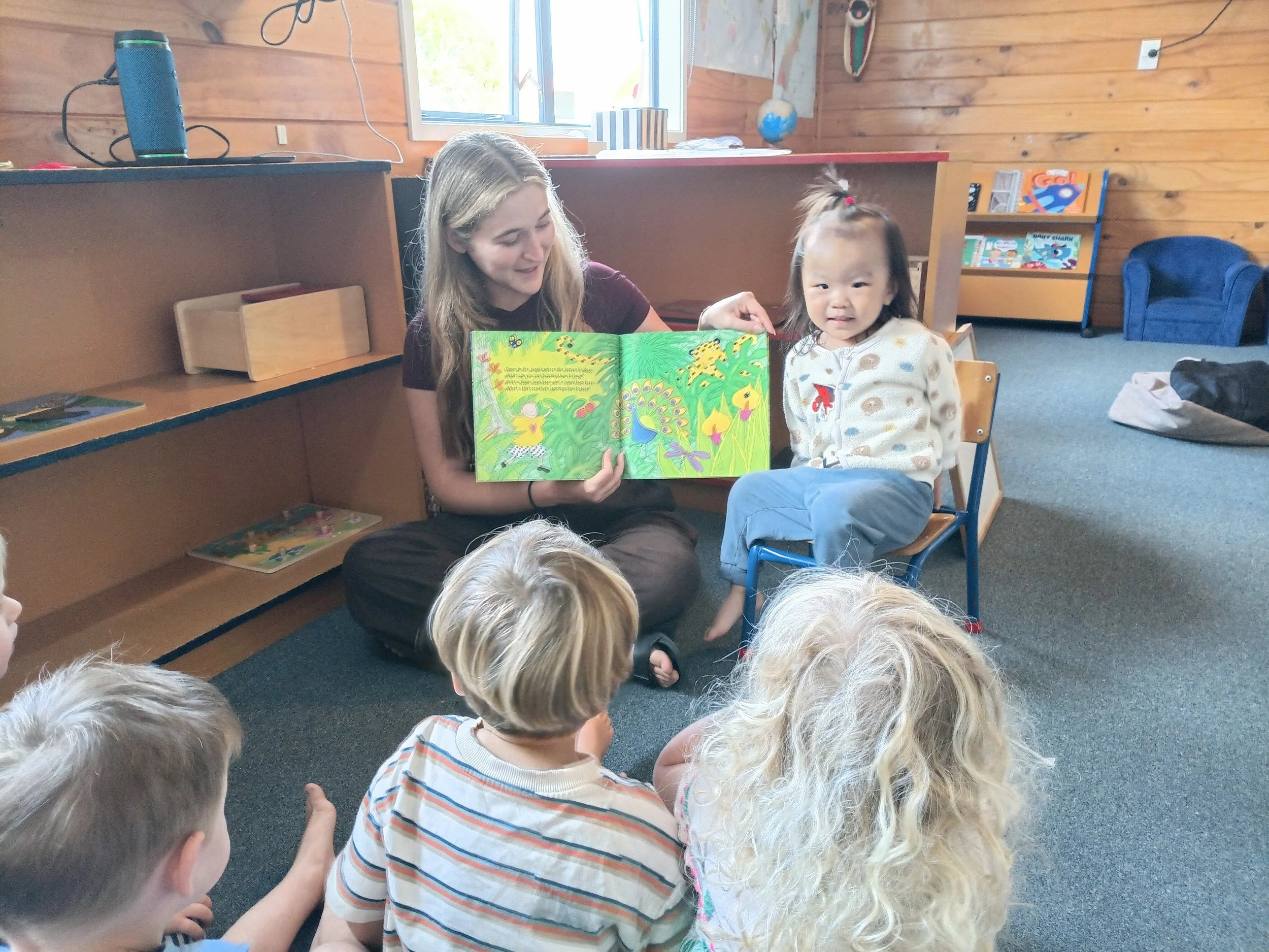 A woman reading a colorful children's book to a group of children in a cozy room with wood-paneled walls.