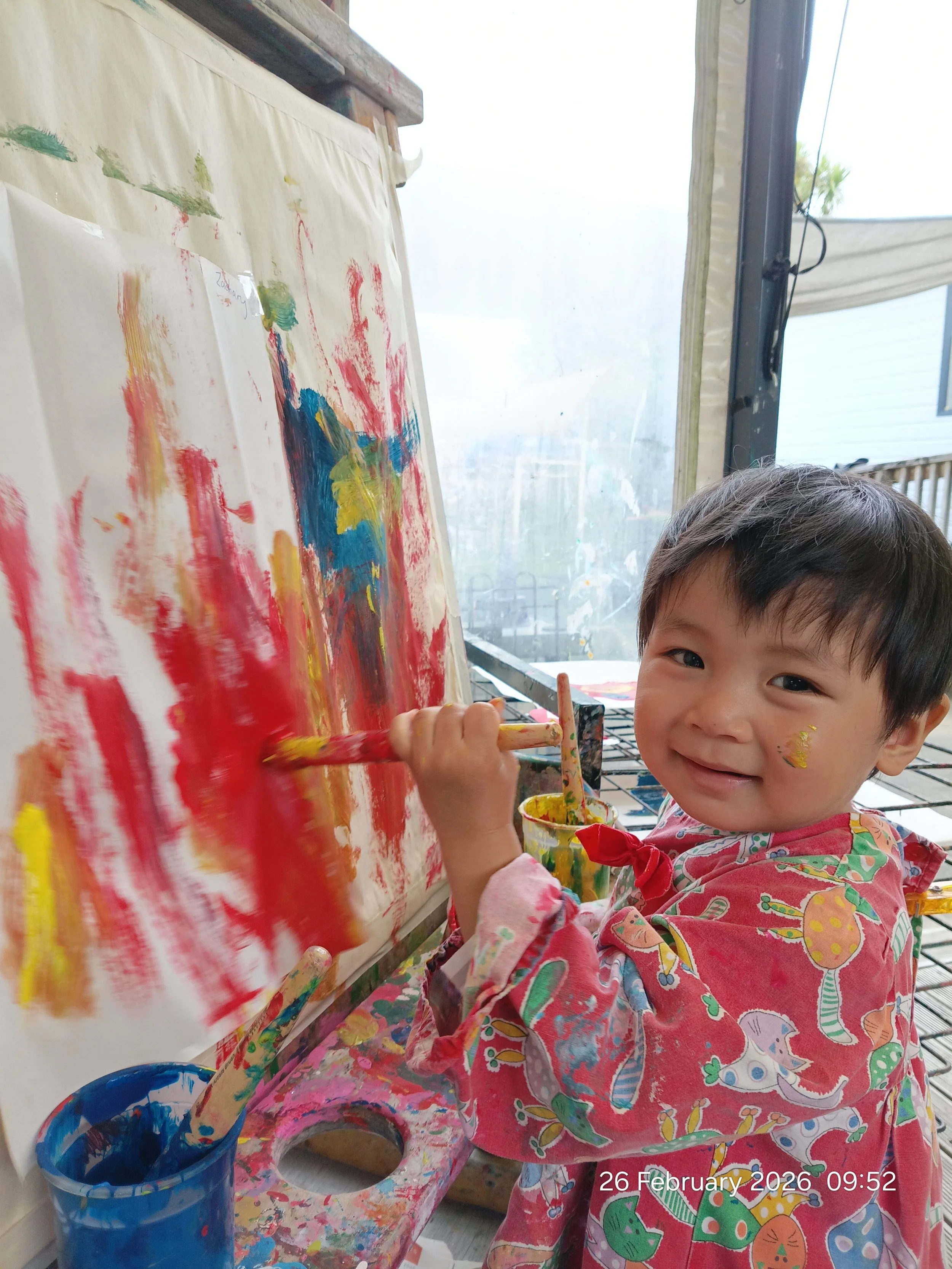 A young child with a smile, wearing a colorful apron, is painting with bright colors on a large canvas inside a room. The child has some paint on their face and is holding a paintbrush, with various painting supplies nearby.