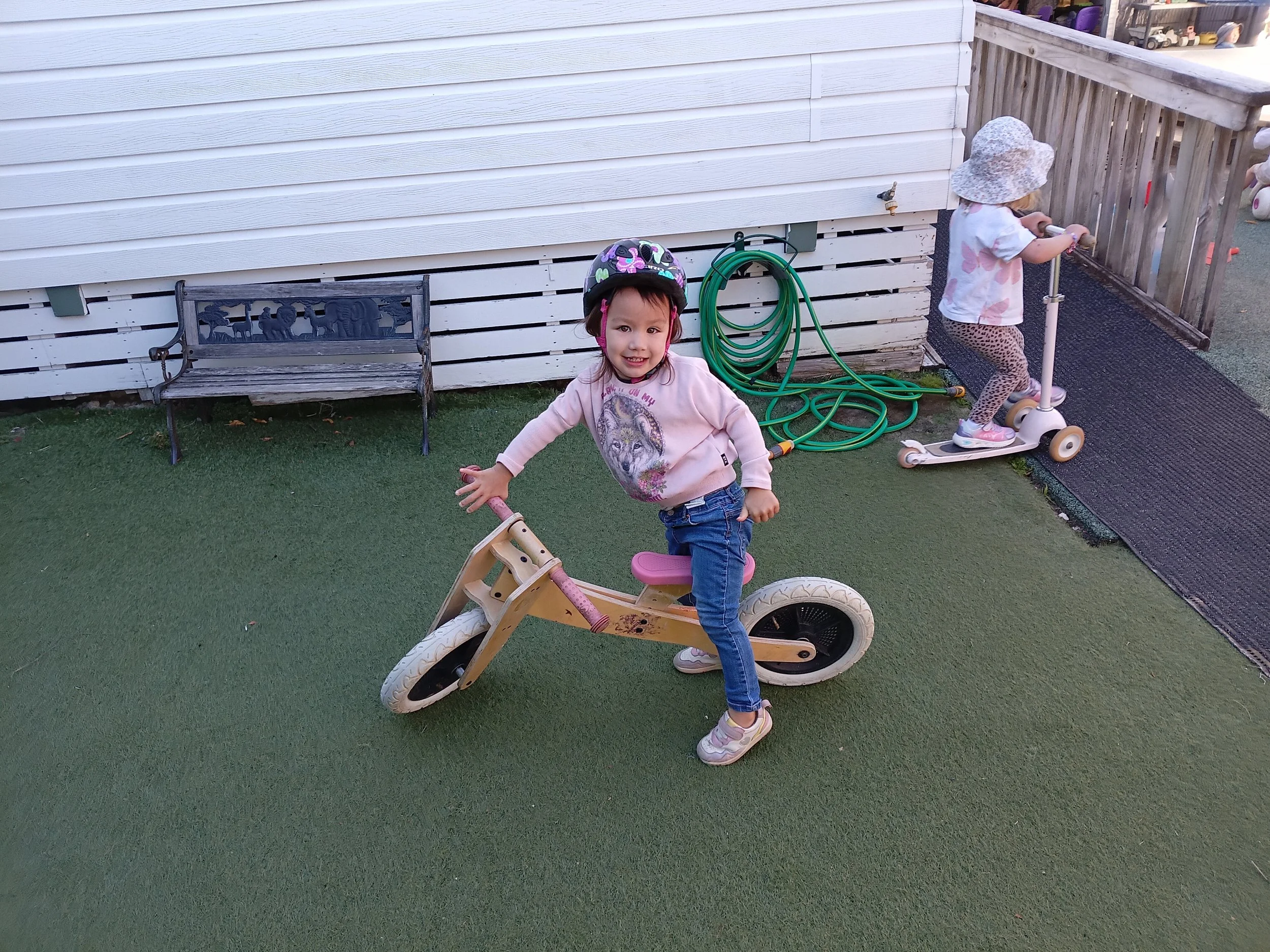 Two young girls playing outside; one girl is sitting on and pretending to ride a pink balance bike with a helmet on, smiling at the camera, and the other girl is riding a scooter near a wooden fence wearing a sun hat.