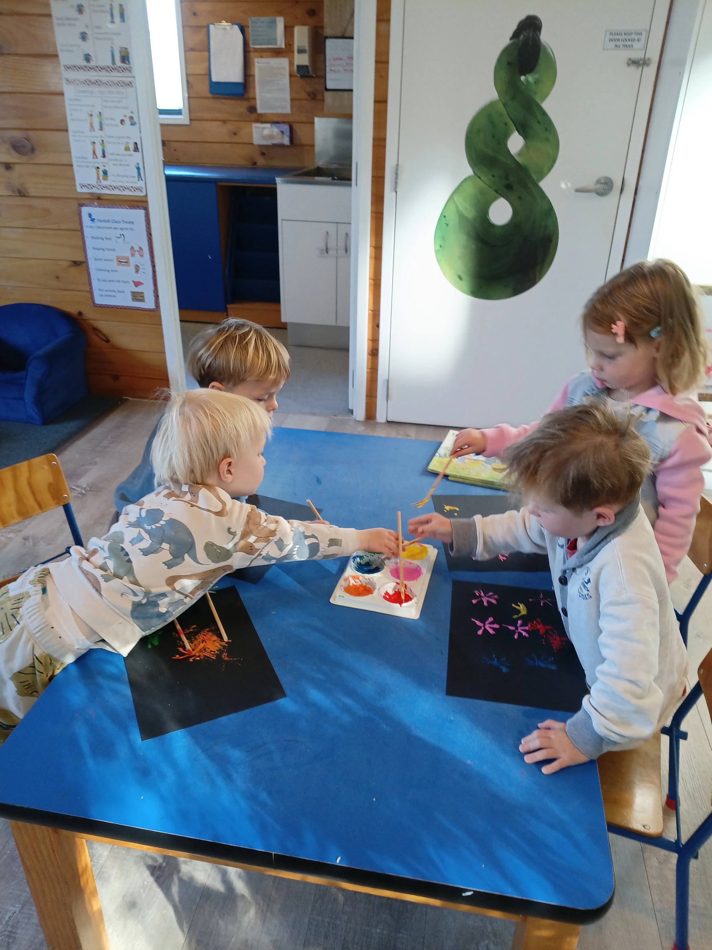 Four children engaged in a painting activity at a blue table, with paint palettes and paper in front of them, in a room with wooden walls and posters.