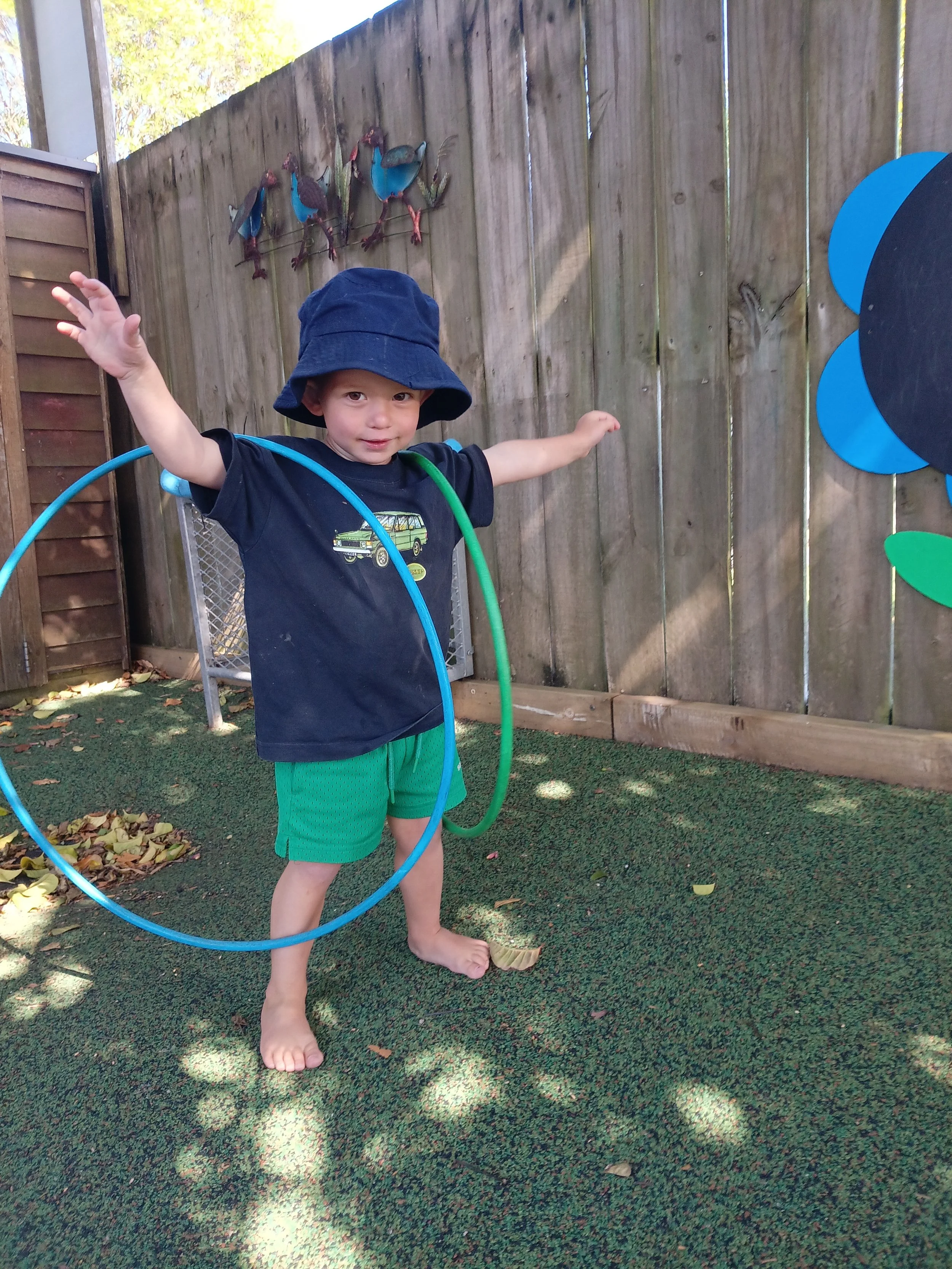 A young child playing outside, wearing a navy bucket hat, a navy t-shirt, green shorts, and barefoot, standing on a green carpeted surface with arms outstretched, surrounded by a blue and green hula hoop, in front of a wooden fence decorated with met