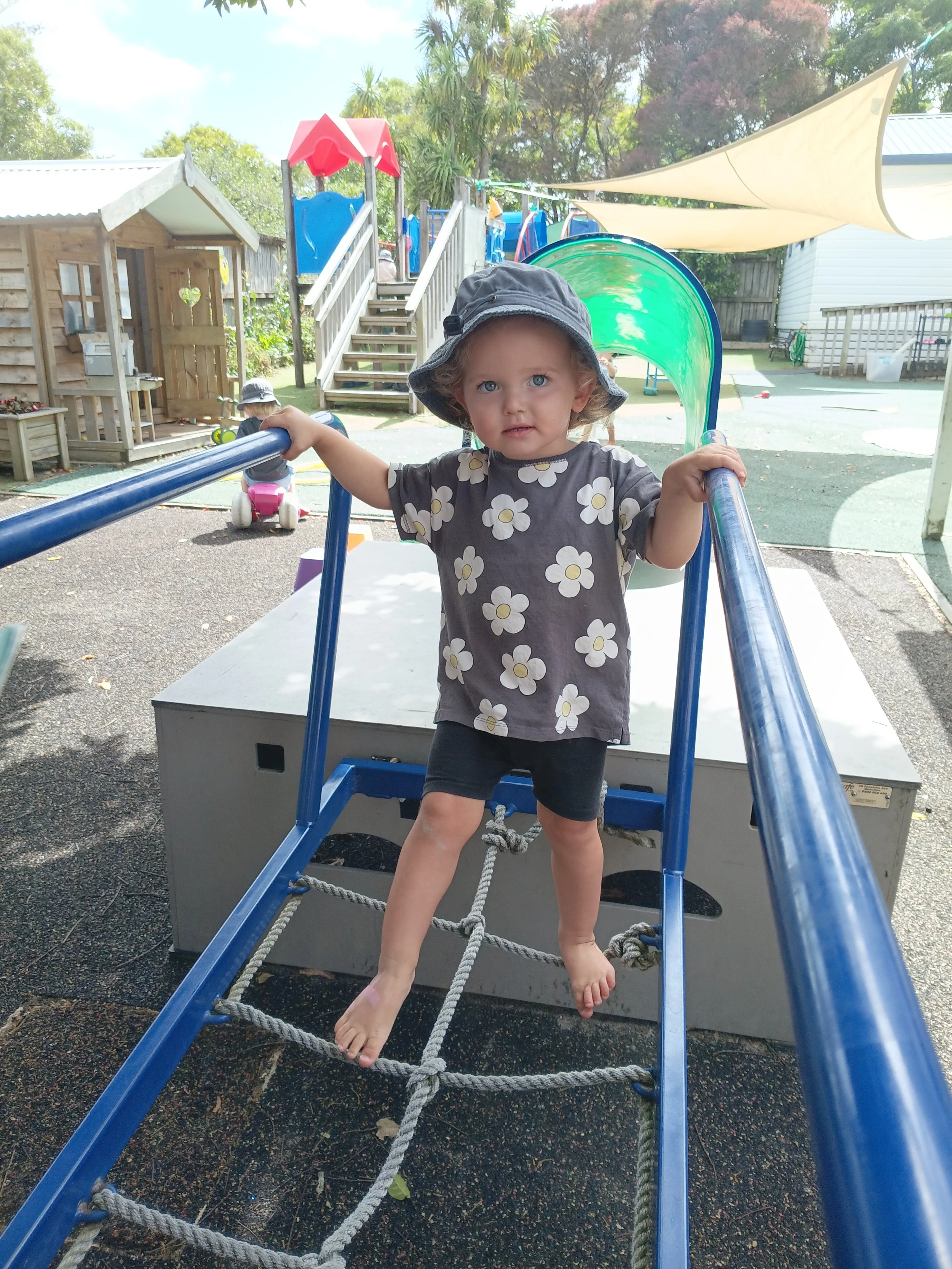 A young girl with curly hair, wearing a gray hat and a gray shirt with white flowers, stands on a play structure in a playground. She is holding onto two blue bars and standing barefoot on a rope ladder. In the background, there are children playing,