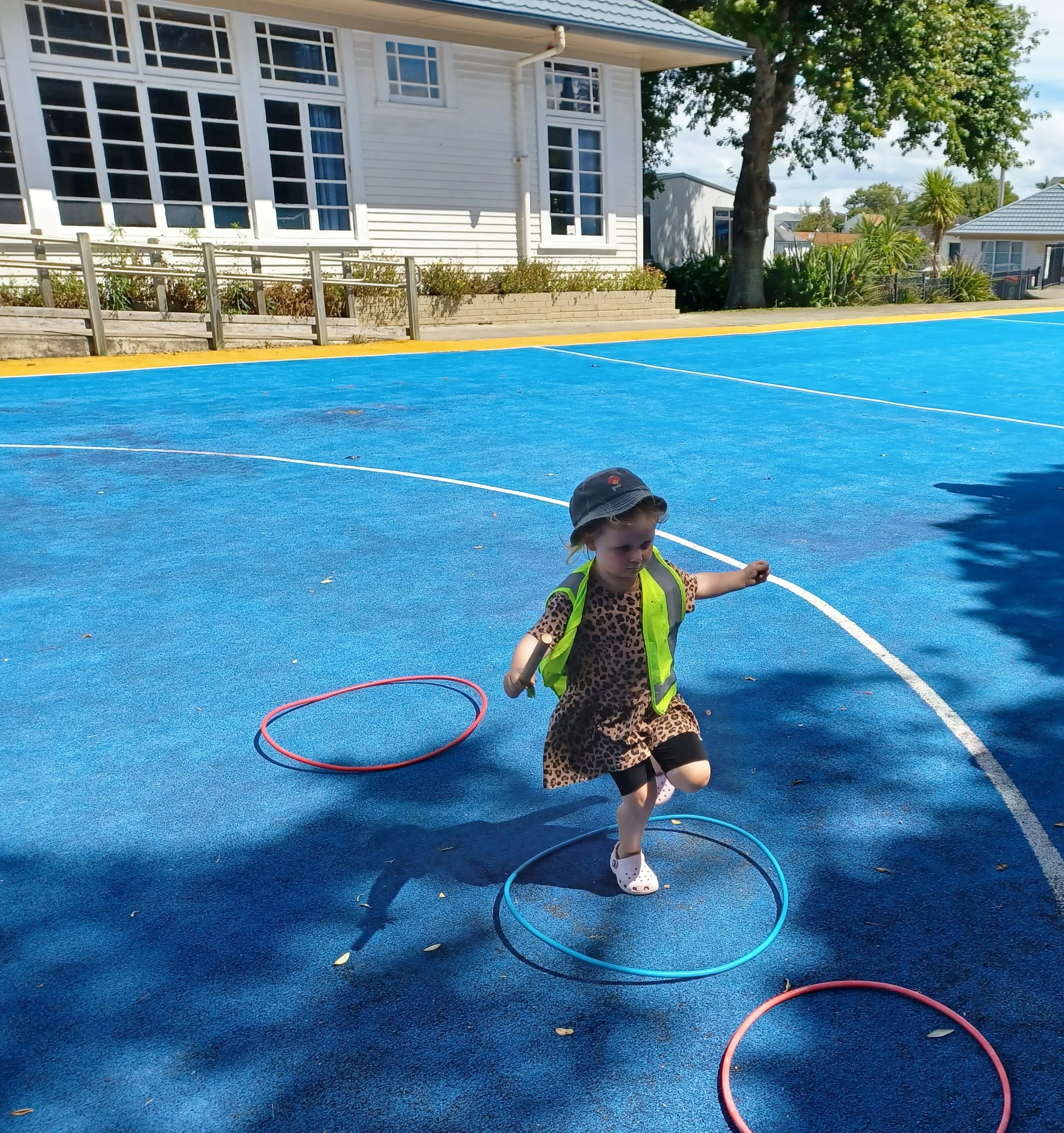 A young girl wearing a leopard print dress, a yellow safety vest, white Crocs, and a bucket hat is playing on a blue playground surface with hula hoops.