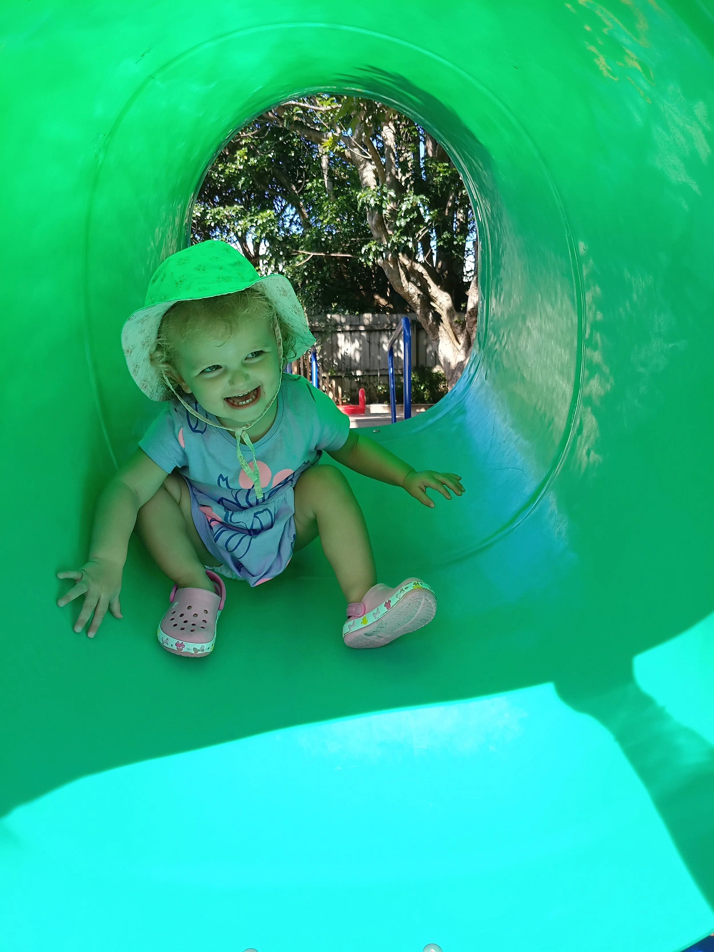 Child crawling inside a green playground tunnel, smiling, wearing a sun hat and pink shoes, outdoors with trees in the background.