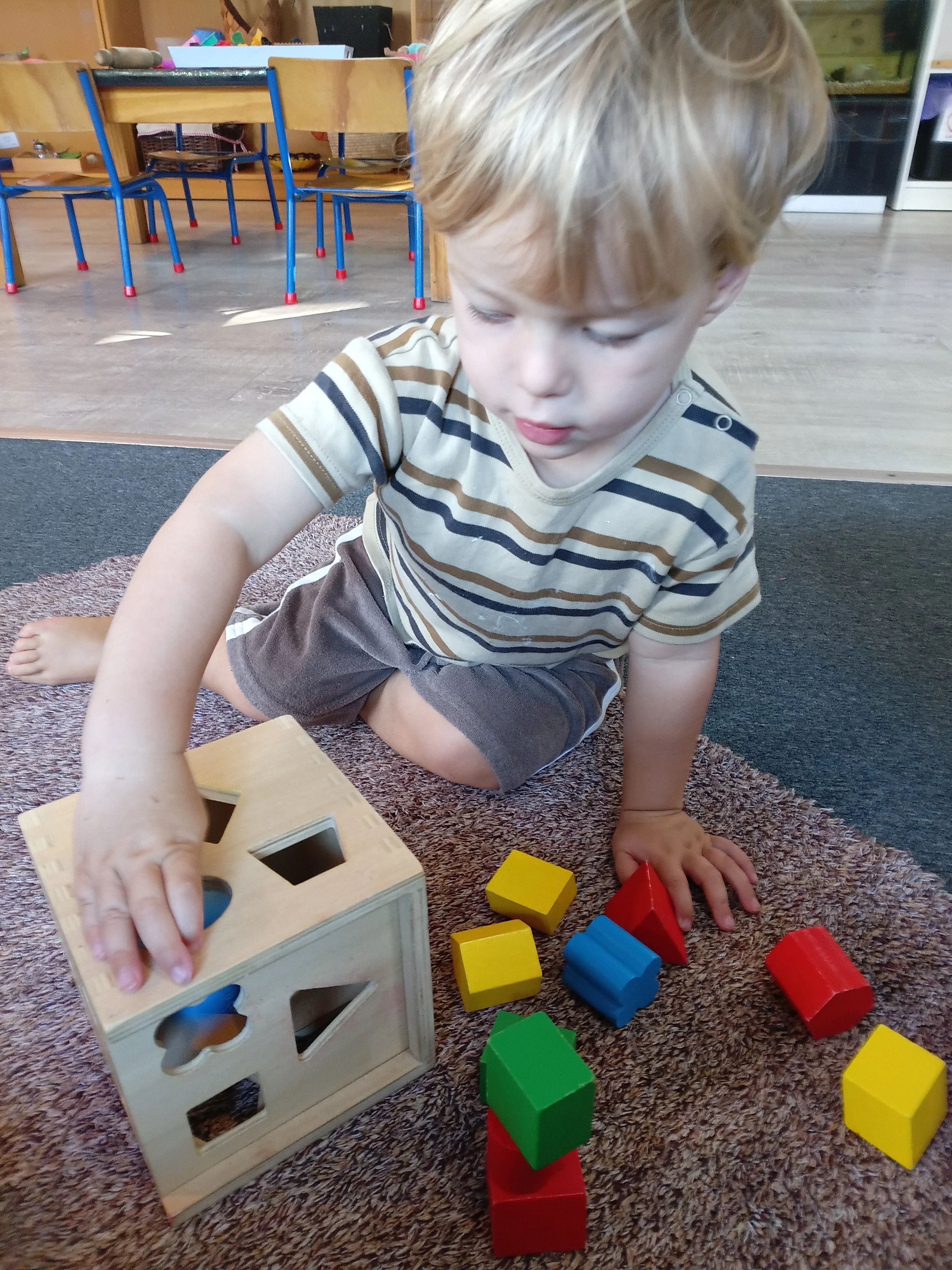 A young boy with blond hair, wearing a striped t-shirt and shorts, playing with colorful wooden blocks on a carpeted floor with a classroom background.