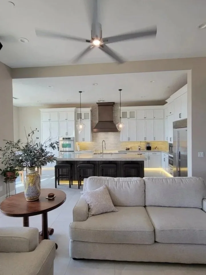 Open-concept living room and kitchen with white cabinetry, a kitchen island with dark wood base, pendant lights, stainless steel appliances, and a ceiling fan.