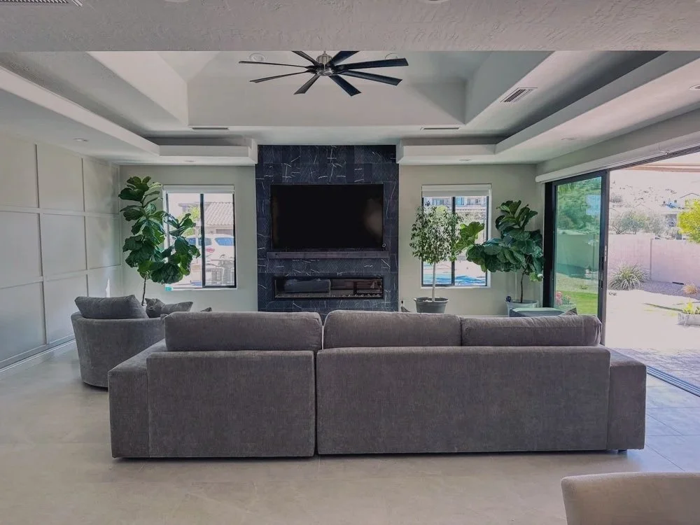 Modern living room with a gray sectional sofa, two armchairs, a black TV mounted above a dark marble fireplace, large potted plants, windows, and a sliding glass door leading to a backyard.