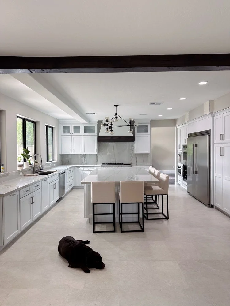 Modern white kitchen with marble countertops, a large island with seating, stainless steel appliances, and a black dog lying on the light-colored floor.