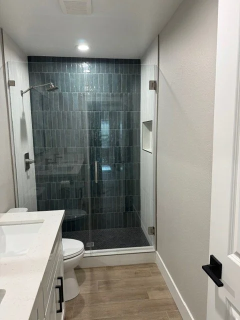 Modern bathroom with glass walk-in shower, white vanity with black handles, and wood-look flooring.