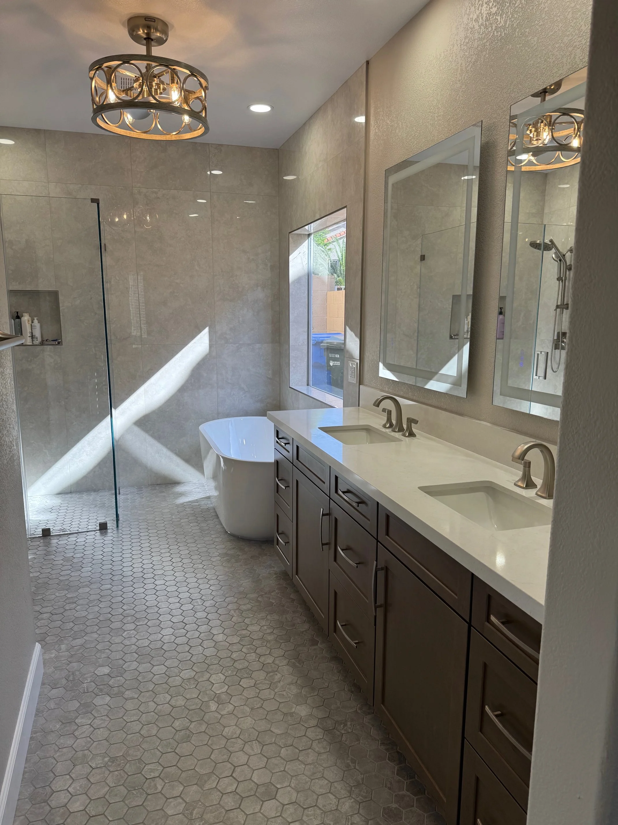 A modern bathroom with a double-sink vanity, large mirror, wall-mounted light fixture, a bathtub, and a shower with glass door. The walls are tiled, and there is a window looking outside.