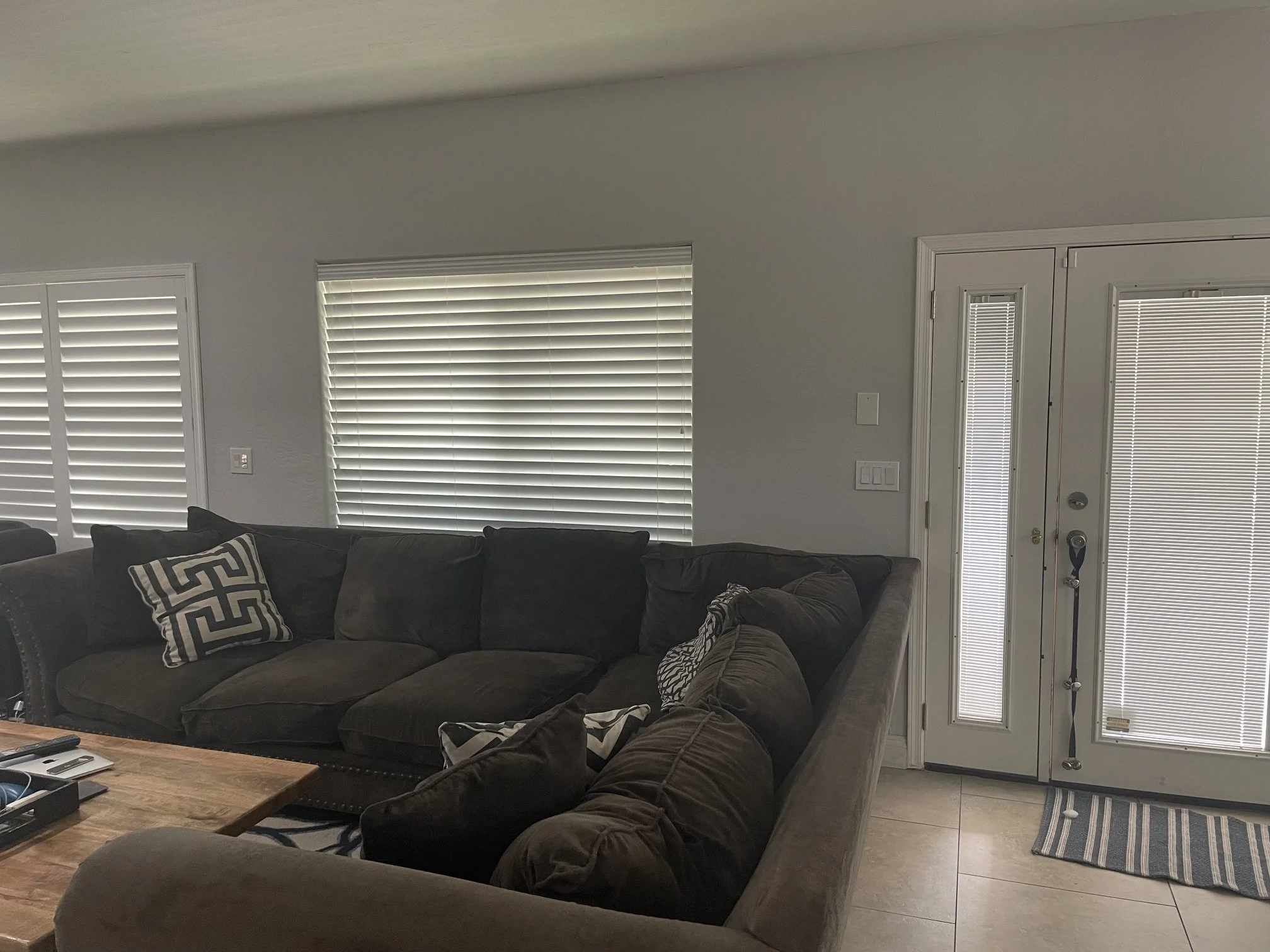 Living room with a large dark brown sectional sofa, patterned pillows, coffee table, and windows with white blinds. There is a door with a narrow window and an outside keypad lock, and a striped rug on the tiled floor.