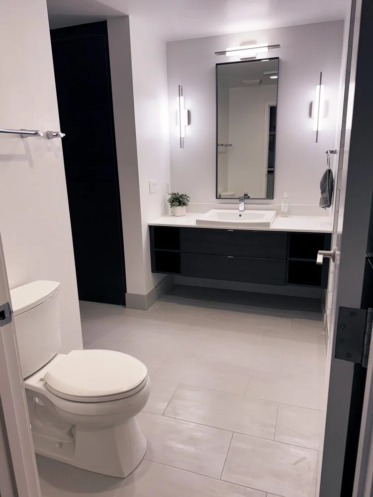 Modern bathroom with a white toilet, a black vanity with a white countertop and sink, a large mirror, and vertical wall lights. The floor is tiled in light gray, and there is a small plant on the vanity.