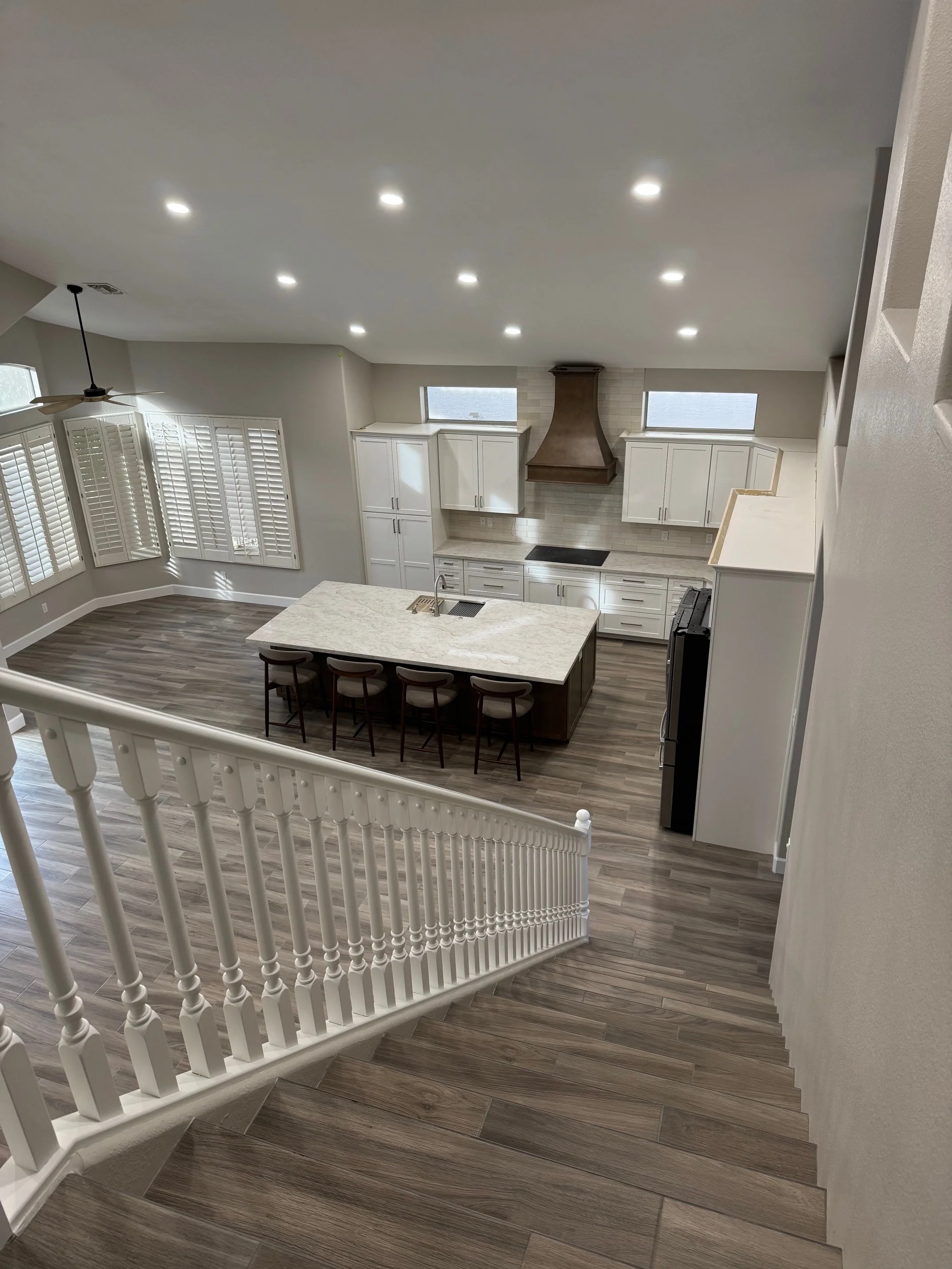 A modern kitchen with white cabinets, a large island with bar stools, a wooden range hood, and hardwood floors. Large windows have white shutters, and recessed ceiling lights illuminate the space.