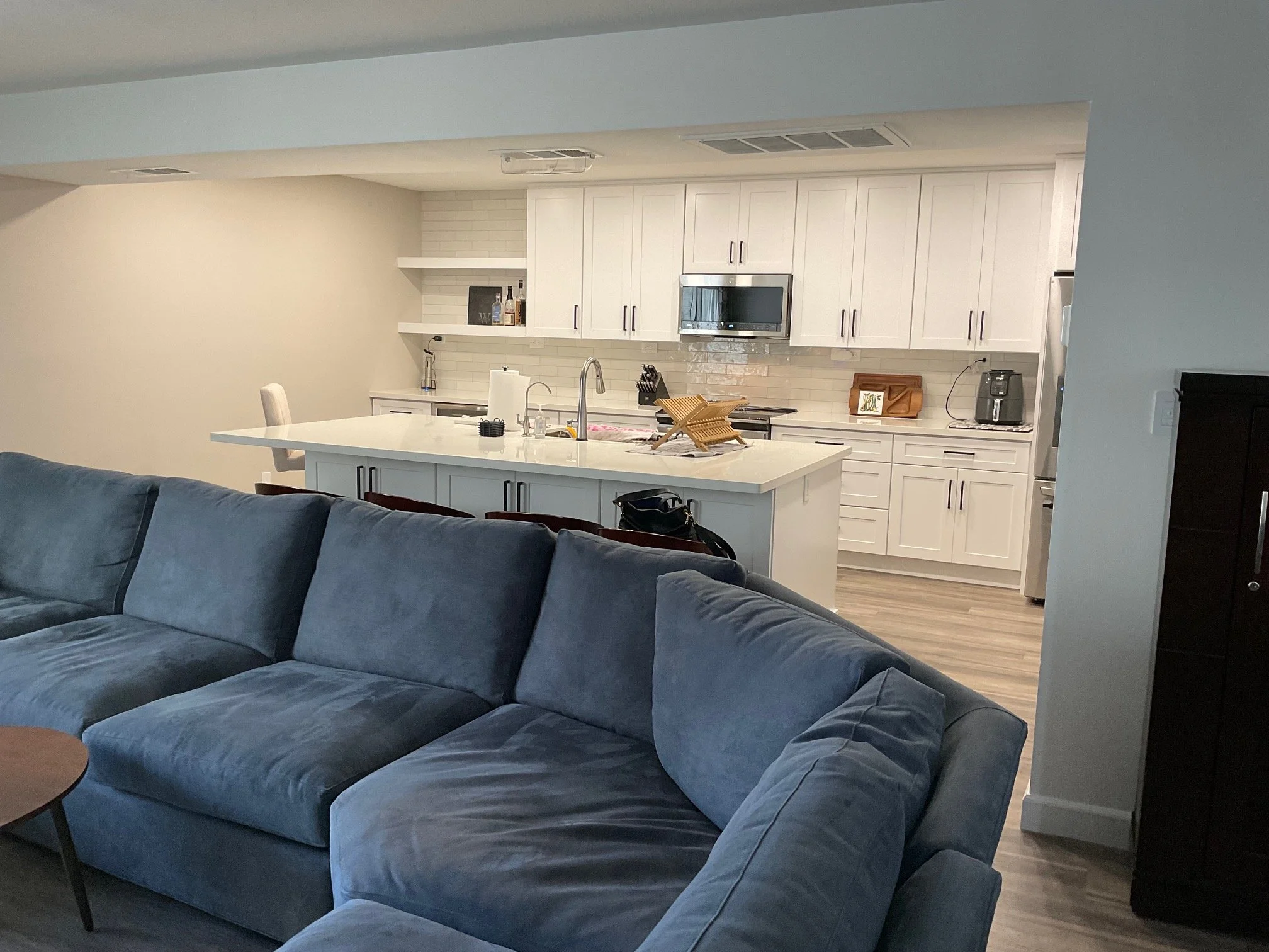 A modern open-concept kitchen with white cabinetry, stainless steel appliances, and a white island with a sink, adjacent to a living room with a dark blue velvet couch.