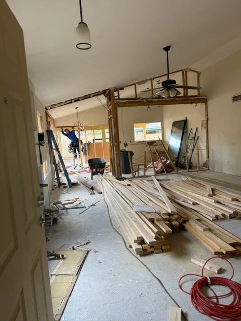 Interior of a house under renovation with exposed framing, construction materials, tools, and a worker in the background.