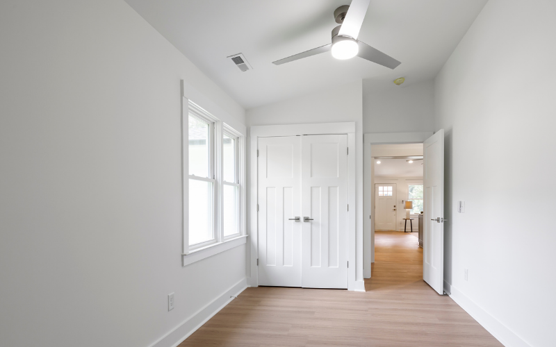 Empty room with white walls, two windows, a white door, and a ceiling fan. Viewed from inside the room, looking into another room with hardwood floors and furniture.