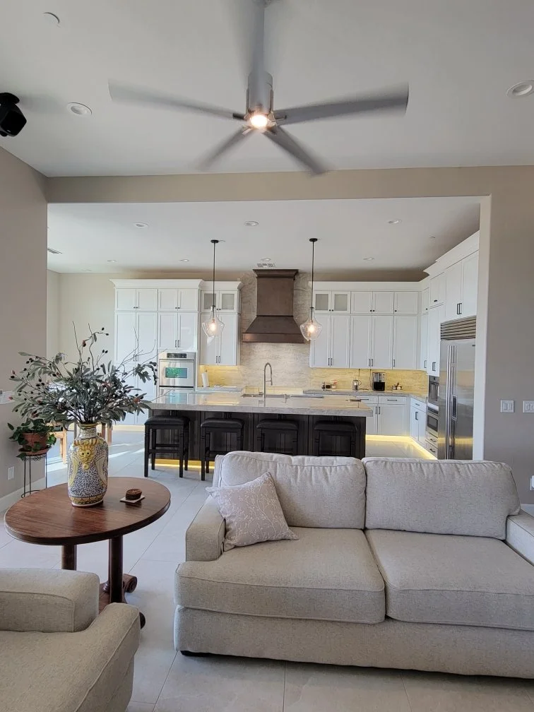 Open-concept living room and kitchen with white cabinets, a kitchen island with black bar stools, and a beige sofa.