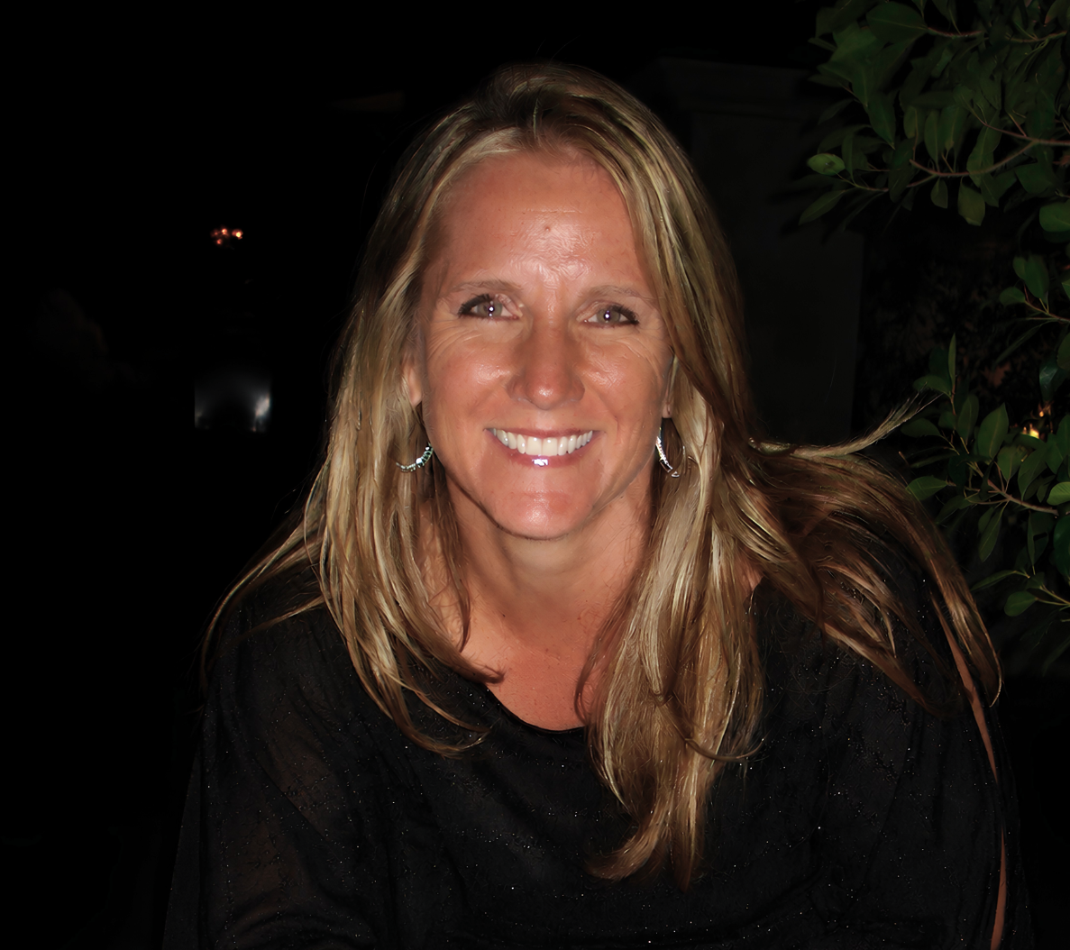 A woman with blonde hair and earrings smiling at the camera, with dark background and green leaves on the right.