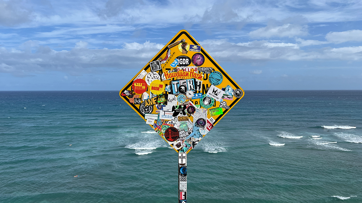 Diamond-shaped sign covered in various stickers, mounted on a pole over the ocean with a background of blue sky and clouds.