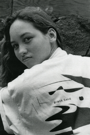 A young woman with long hair looking at the camera, outside with rocks in the background, wearing a casual shirt.