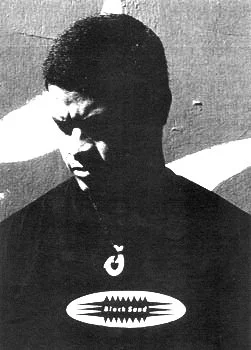 Black and white image of a young man with short hair, wearing a dark jacket, standing in front of a graffiti-covered wall.