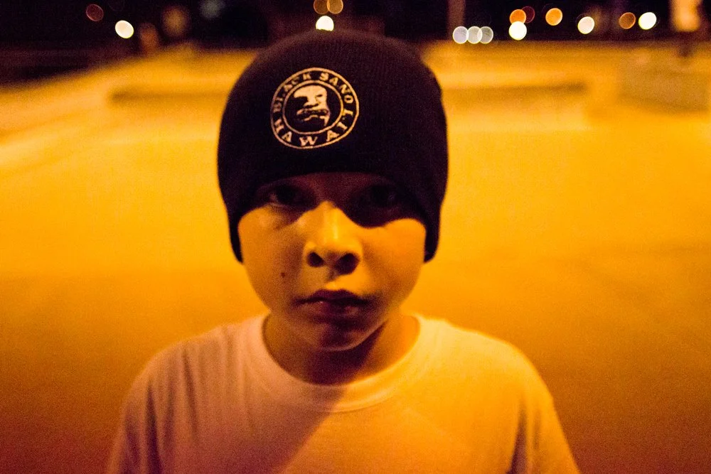 Young person wearing a black beanie with a white logo, outdoors at night with blurred streetlights in the background.