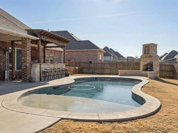 Backyard with in-ground pool, outdoor fireplace, and a covered patio area with bar stools, surrounded by a wooden fence and neighboring houses.