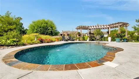 A backyard swimming pool with a stone border, surrounded by a concrete patio, green bushes, trees, and a wooden pergola under a blue sky.