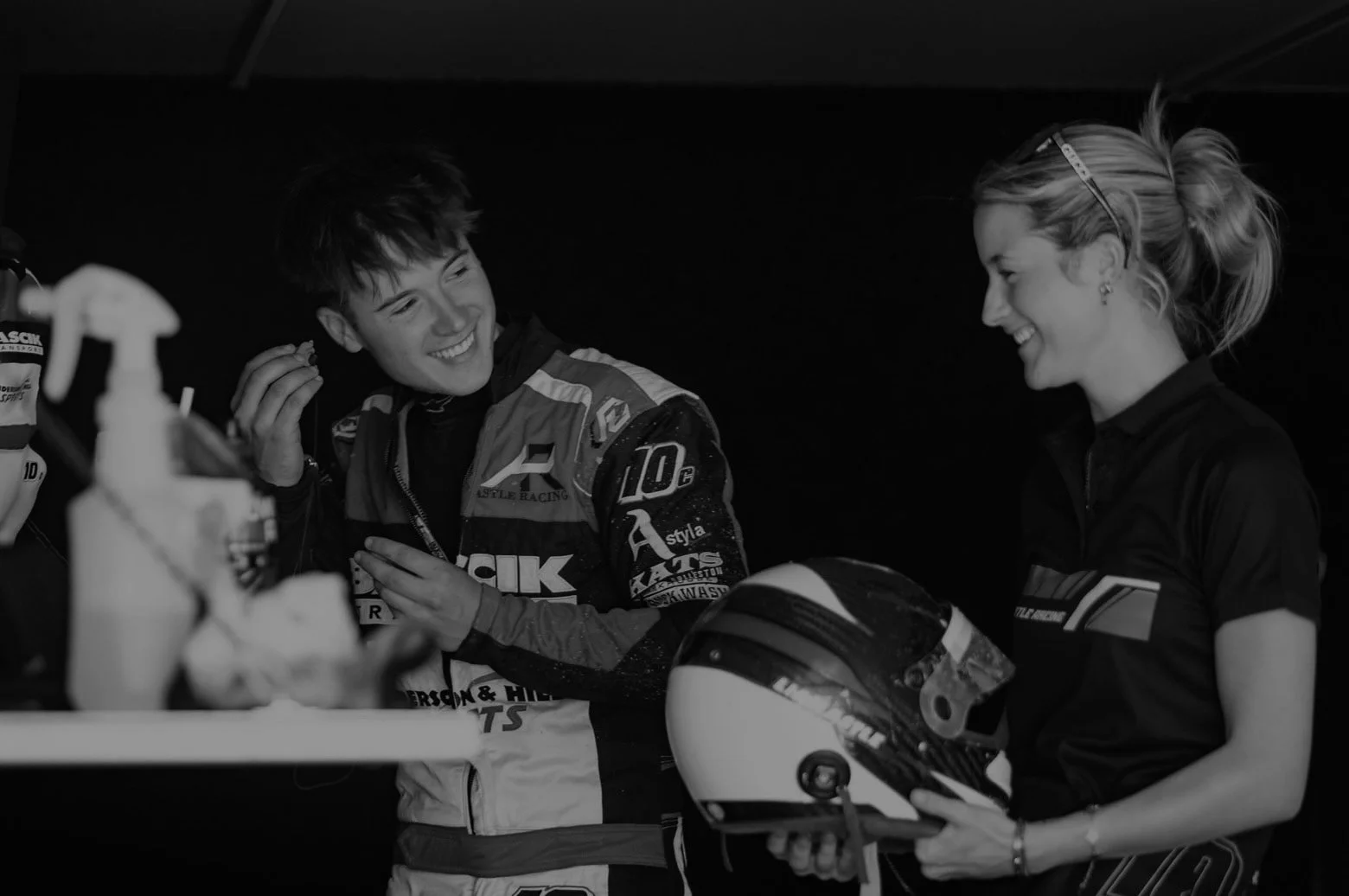 Two smiling women and a man in racing gear inside a garage, with a helmet and bottles on a table.