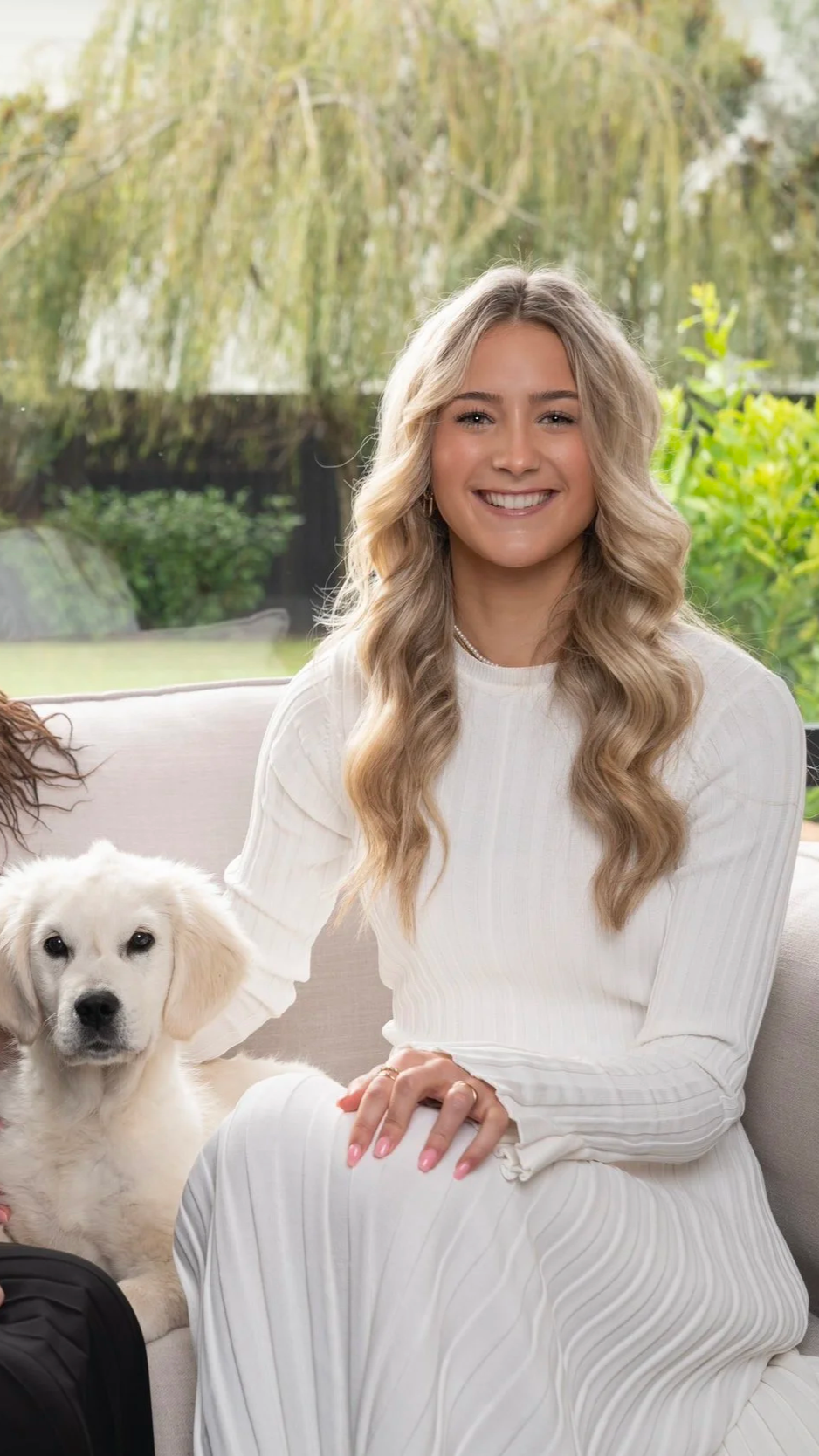 A young woman with long, wavy blonde hair, smiling, sitting on a beige couch with a white puppy beside her, in front of large window with a garden view.