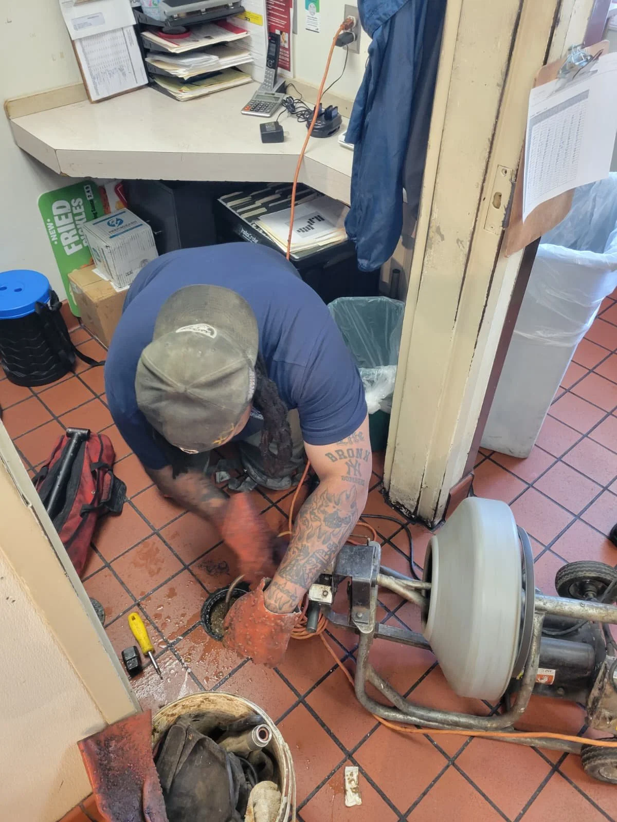 A worker using specialized equipment to extract a drain clog in a restaurant kitchen, with various tools and supplies nearby.